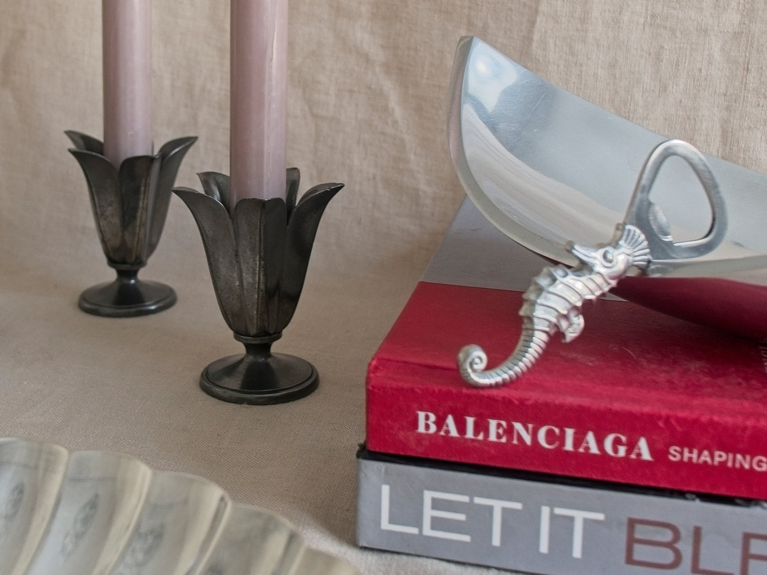A pair of Just andersen Danish pewter art deco 1920s tulip shaped candle holders next to a Just Andersen pewter seahorse shaped bottle opener in front of a neutral linen backdrop