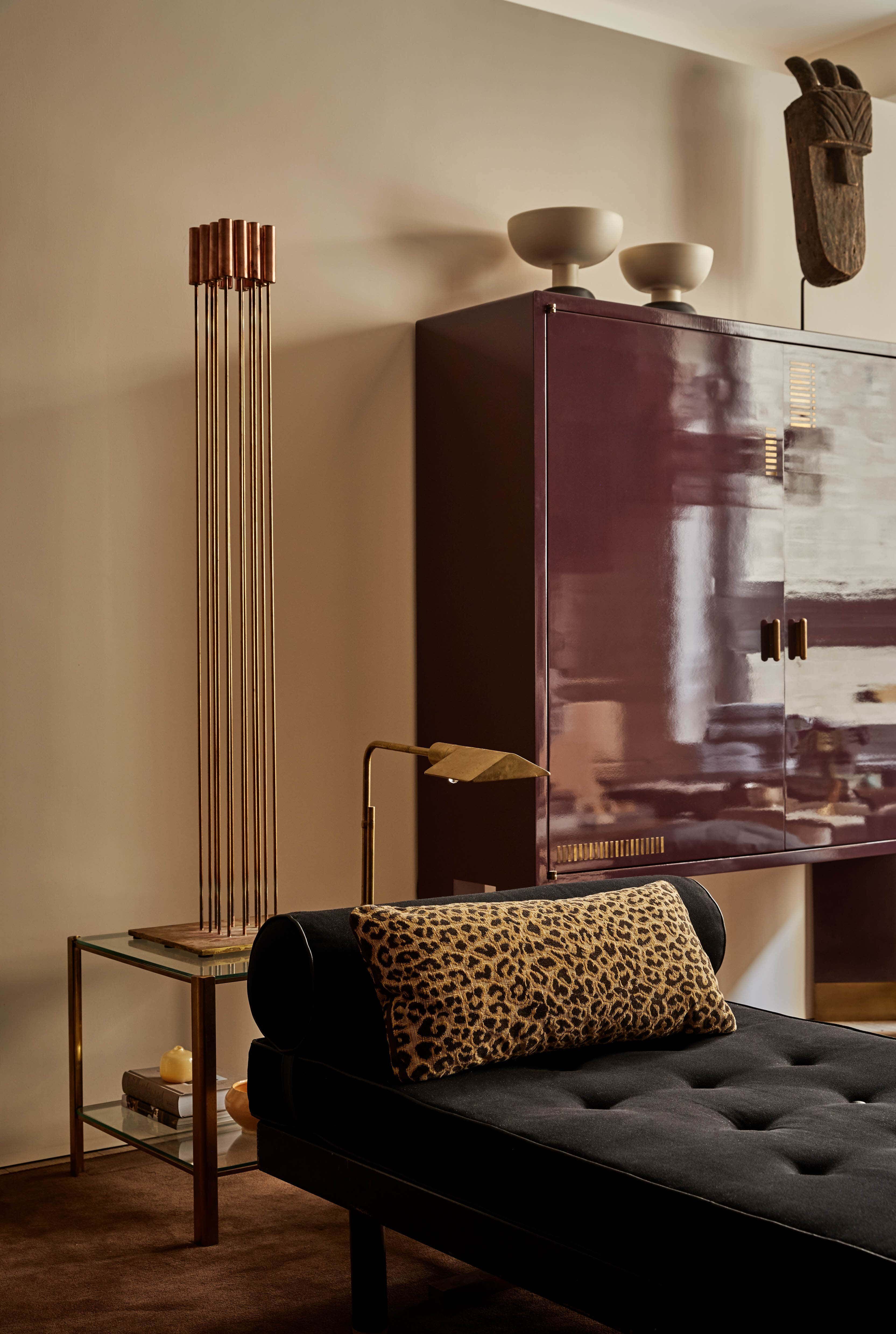 A peach room with a glossy plum coloured cabinet, a black velvet chaise longue with a rectangular leopard print cushion, a small glass and chrome mid century modern side table and a vintage 1970s art deco gold floor lamp