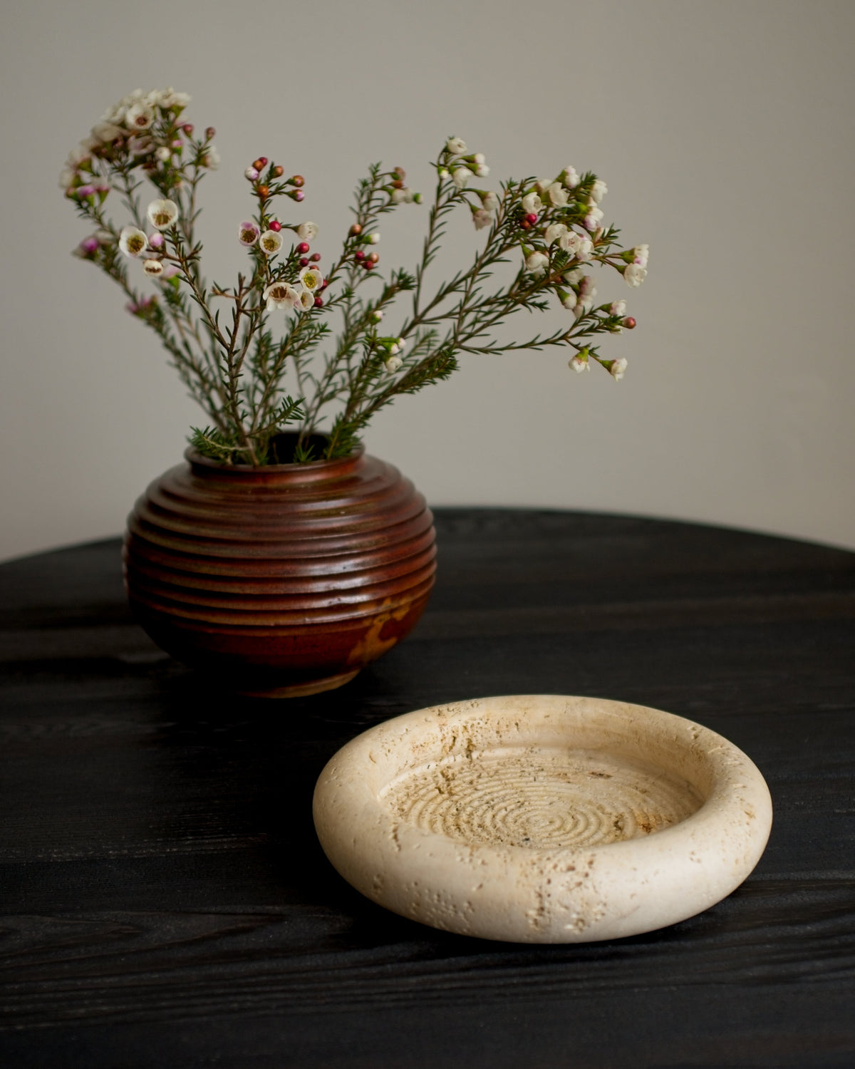 A vintage mid century modern round ceramic ridged vase containing small white flowers behind a Fratelli Mannelli travertine hoe decor piece on a balck wooden surface in front of a grey background.