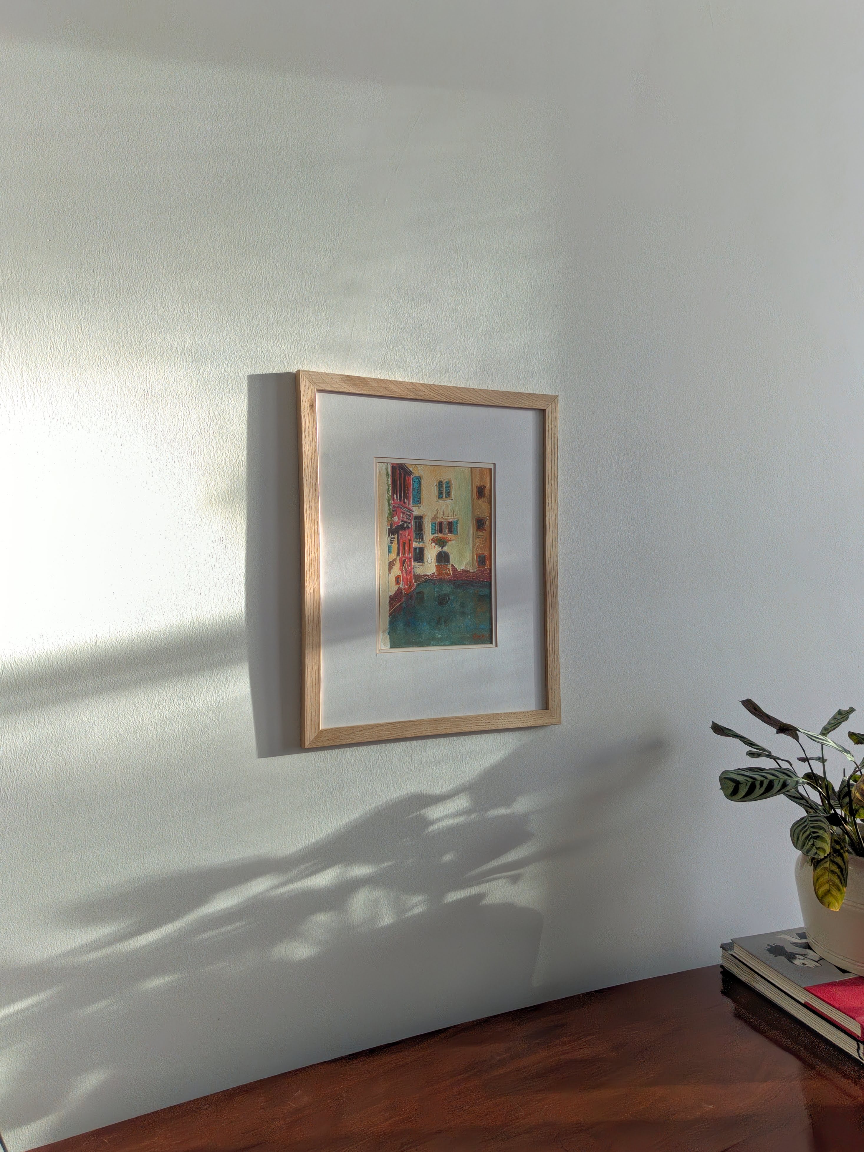A pine framed vintage watercolour painting of a colourful Venetian canal scene on a white wall with a plant to the right.