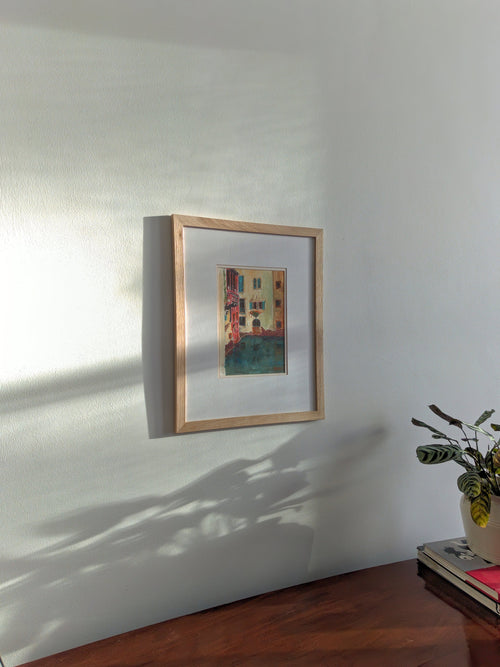 A pine framed vintage watercolour painting of a colourful Venetian canal scene on a white wall with a plant to the right.