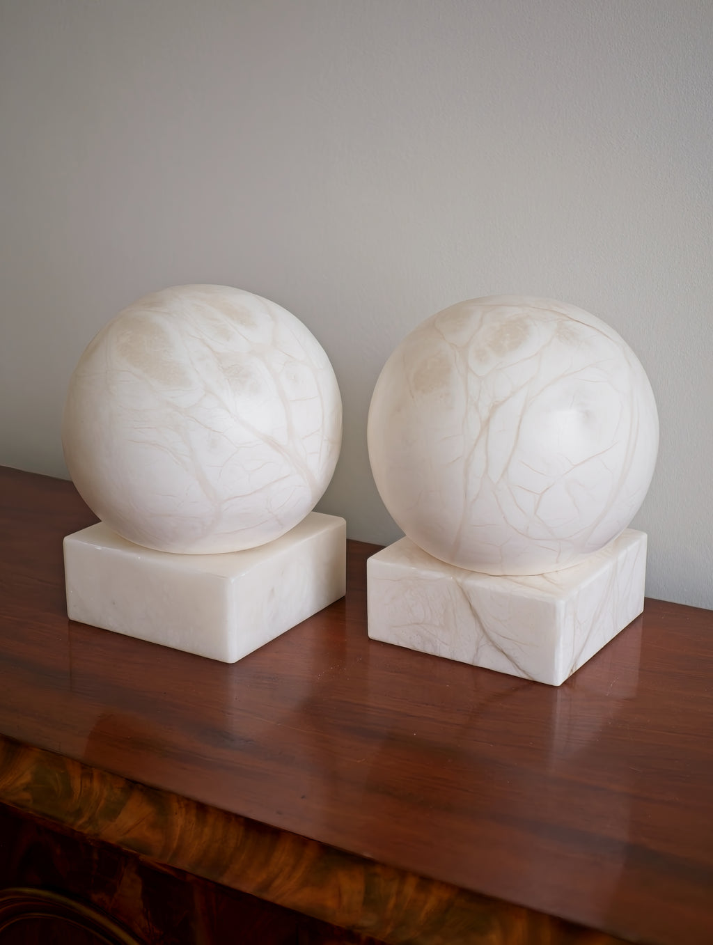 Two white marble spheres on square bases against a plain wall on a brown walnut wood surface.