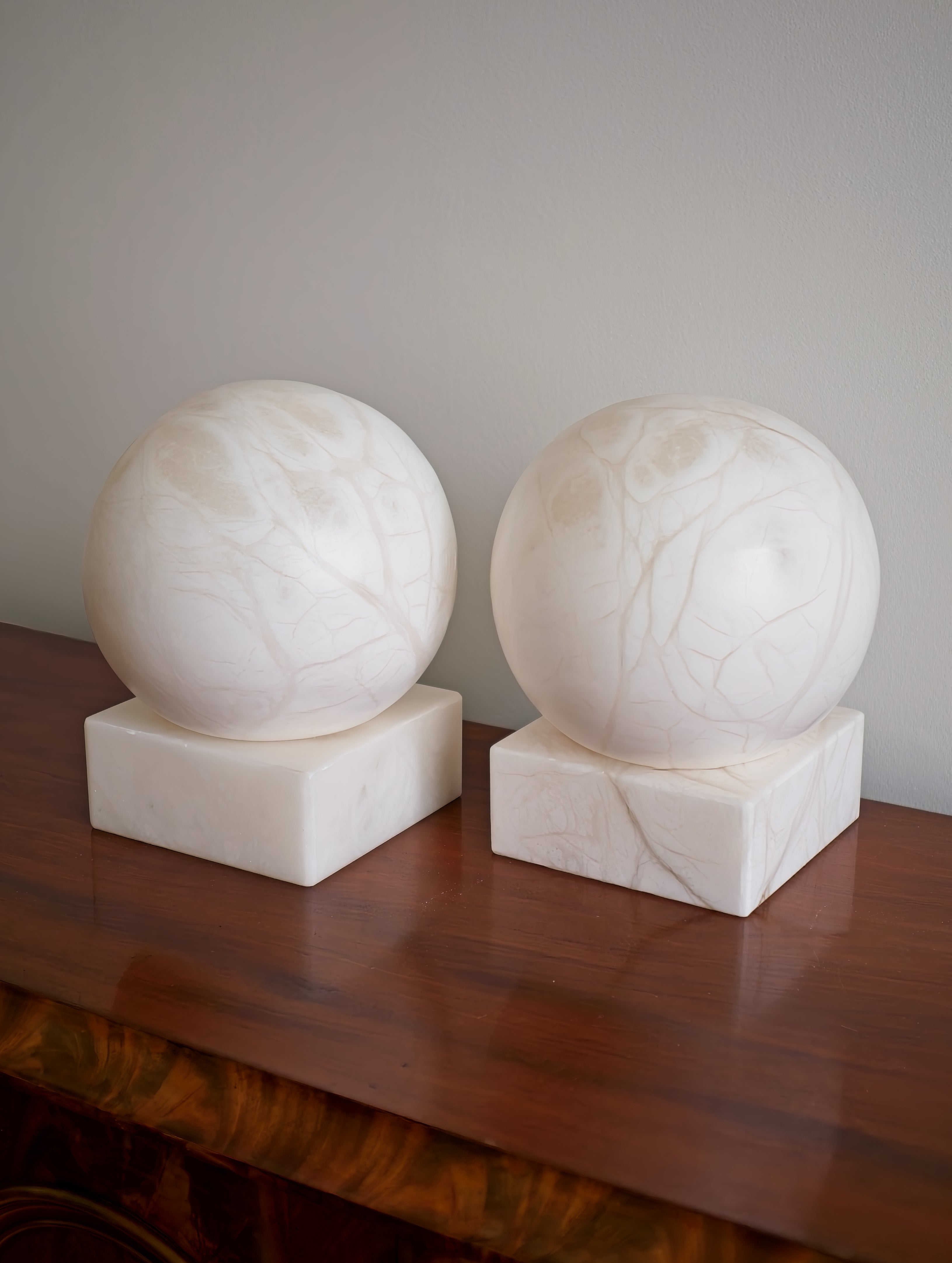 Two white marble spheres on square bases against a plain wall on a brown walnut wood surface.