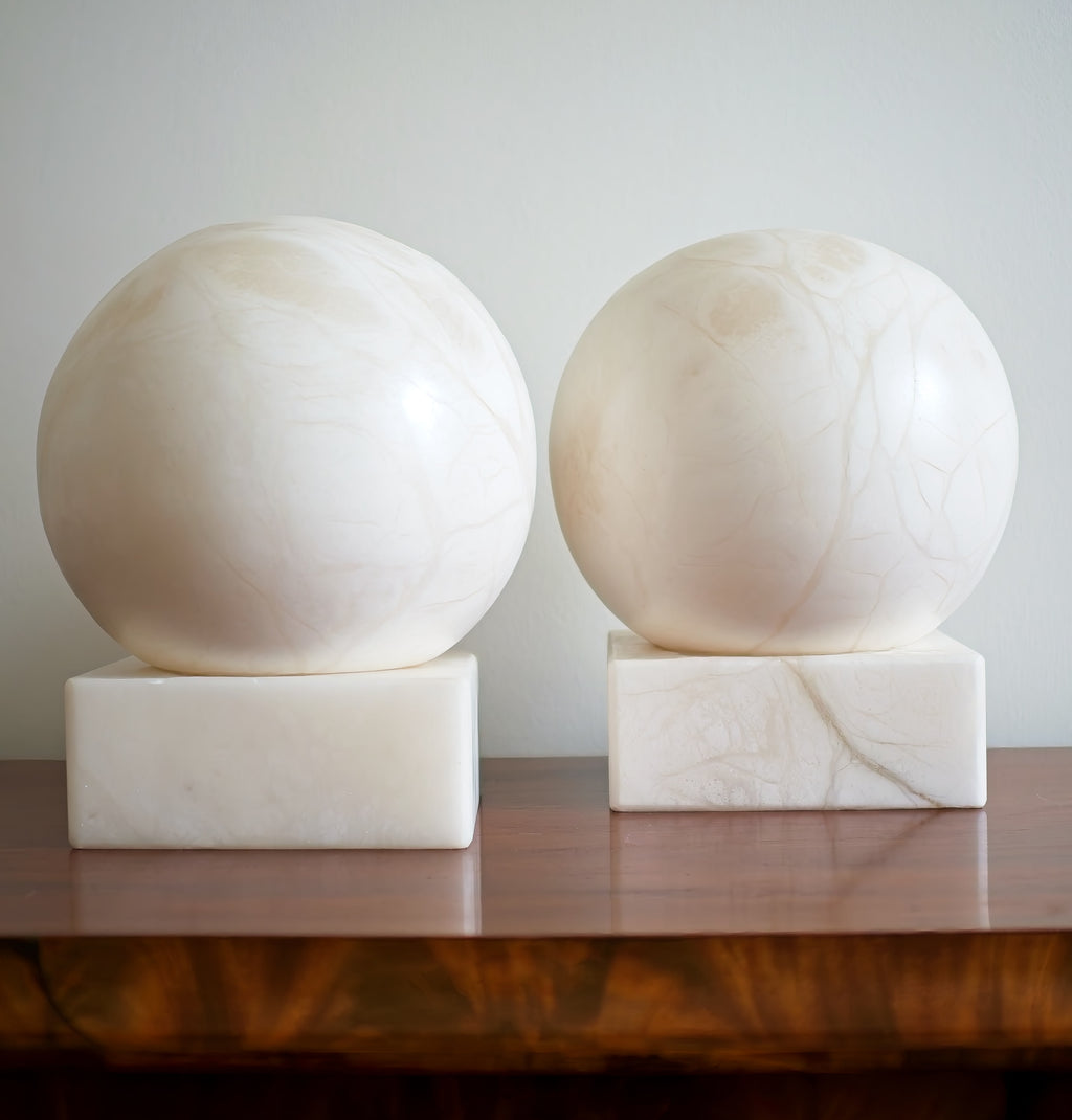 A close up of two mid century modern vintage alabaster marble moon shaped lamps on square bases against a plain background from the front.