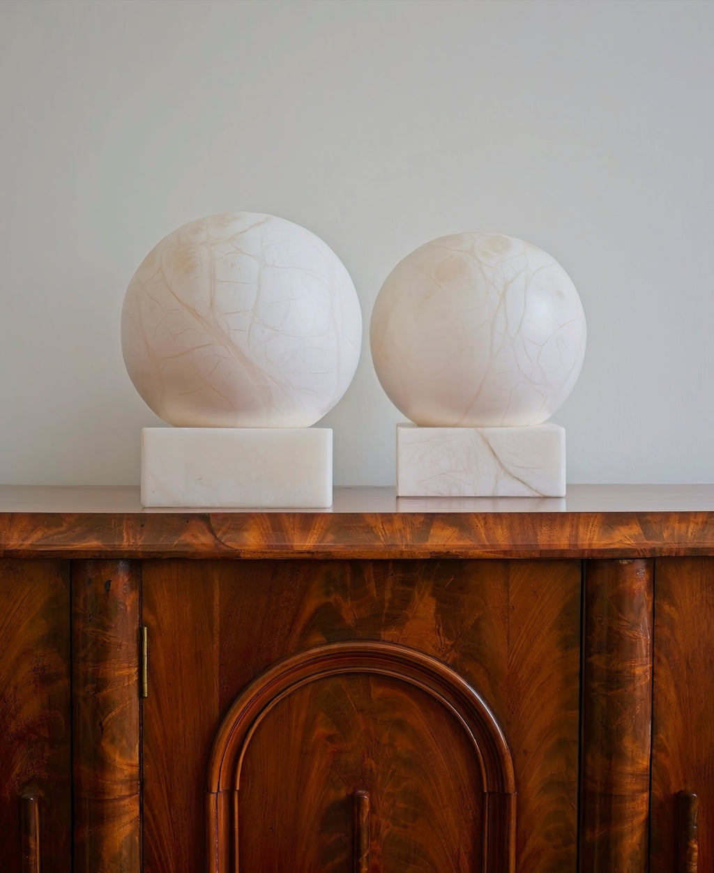 A pair of art deco 1970s mid century modern vintage white alabaster globe shaped table lamps on an antique burr wood cabinet against a white wall.