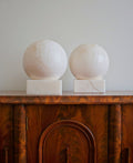 A pair of art deco 1970s mid century modern vintage white alabaster globe shaped table lamps on an antique burr wood cabinet against a white wall.