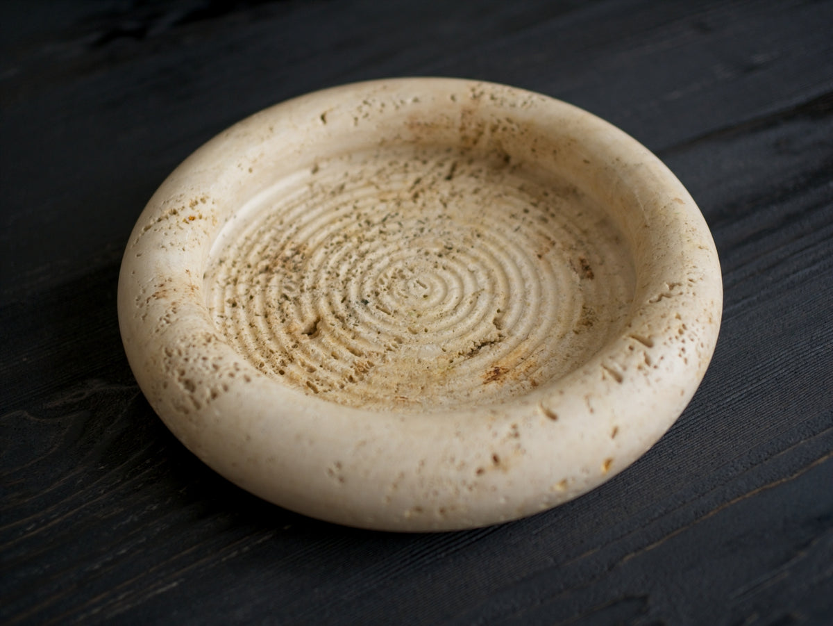 A 1970s vintage Fratelli Mannelli travertine vide poche on a black wood surface. This circular mid century modern travertine dish is hand carved with engraved circular ridge work inside, a wide sculptural rim and crackled texture throughout and is an beautiful example of classic retro Italian home decor that would suit modern vintage, retro, minimalist, Hollywood Regency, modernist or art deco inspired home interior schemes.