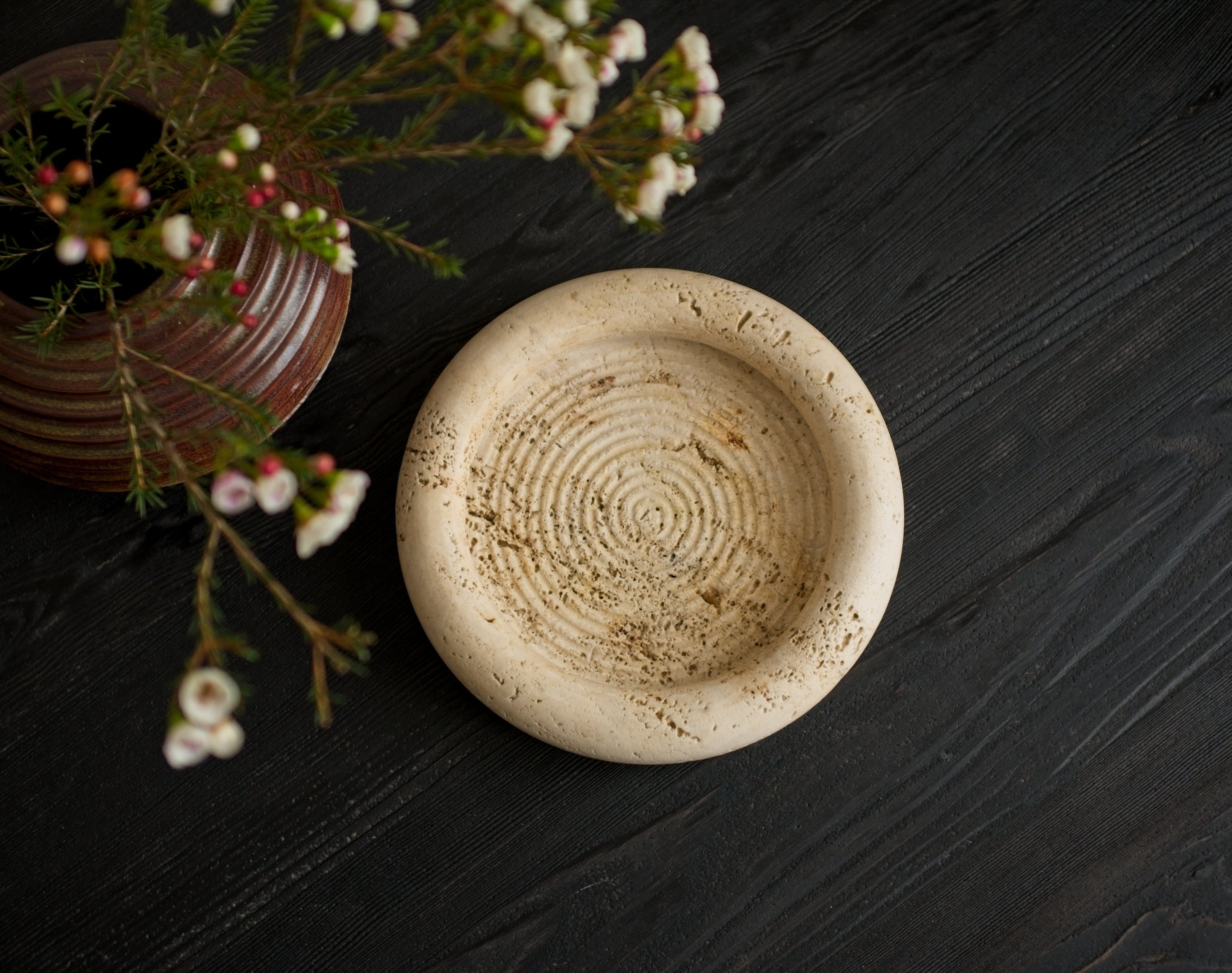 Ceramic vase on a black wooden surface with floral decorations next to a round beige stone dish with circular ridged details inside.