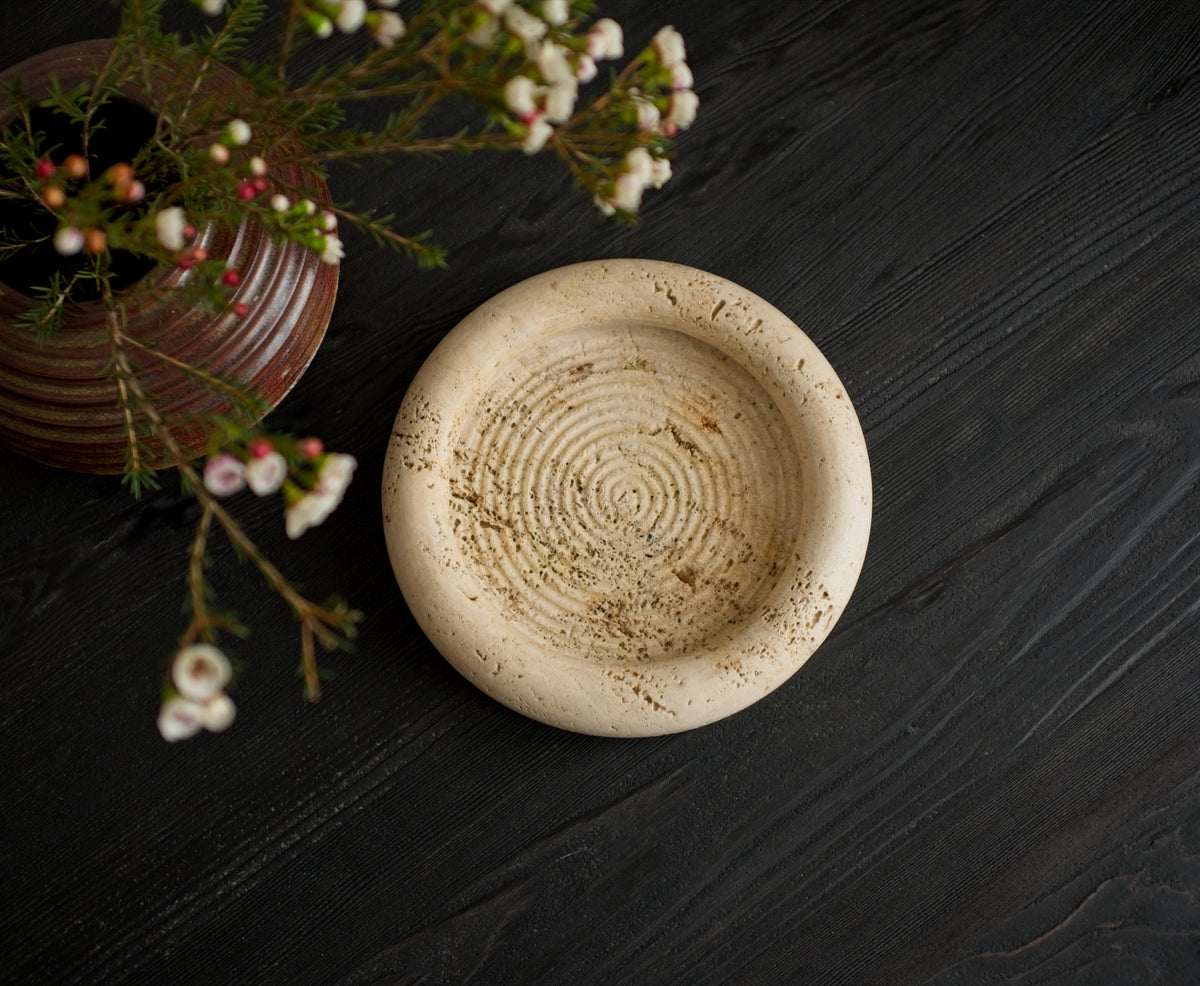 A 1970s vintage Fratelli Mannelli travertine vide poche next to a brutalist globe vase containing small white rustic flowers on a black wood surface shot from above. The mid century modern travertine dish is hand carved with engraved circular ridge work inside, a wide sculptural rim and crackled texture throughout and is an beautiful example of classic retro Italian home decor that would suit modern vintage, retro, minimalist, Hollywood Regency, modernist or art deco inspired home interior schemes.