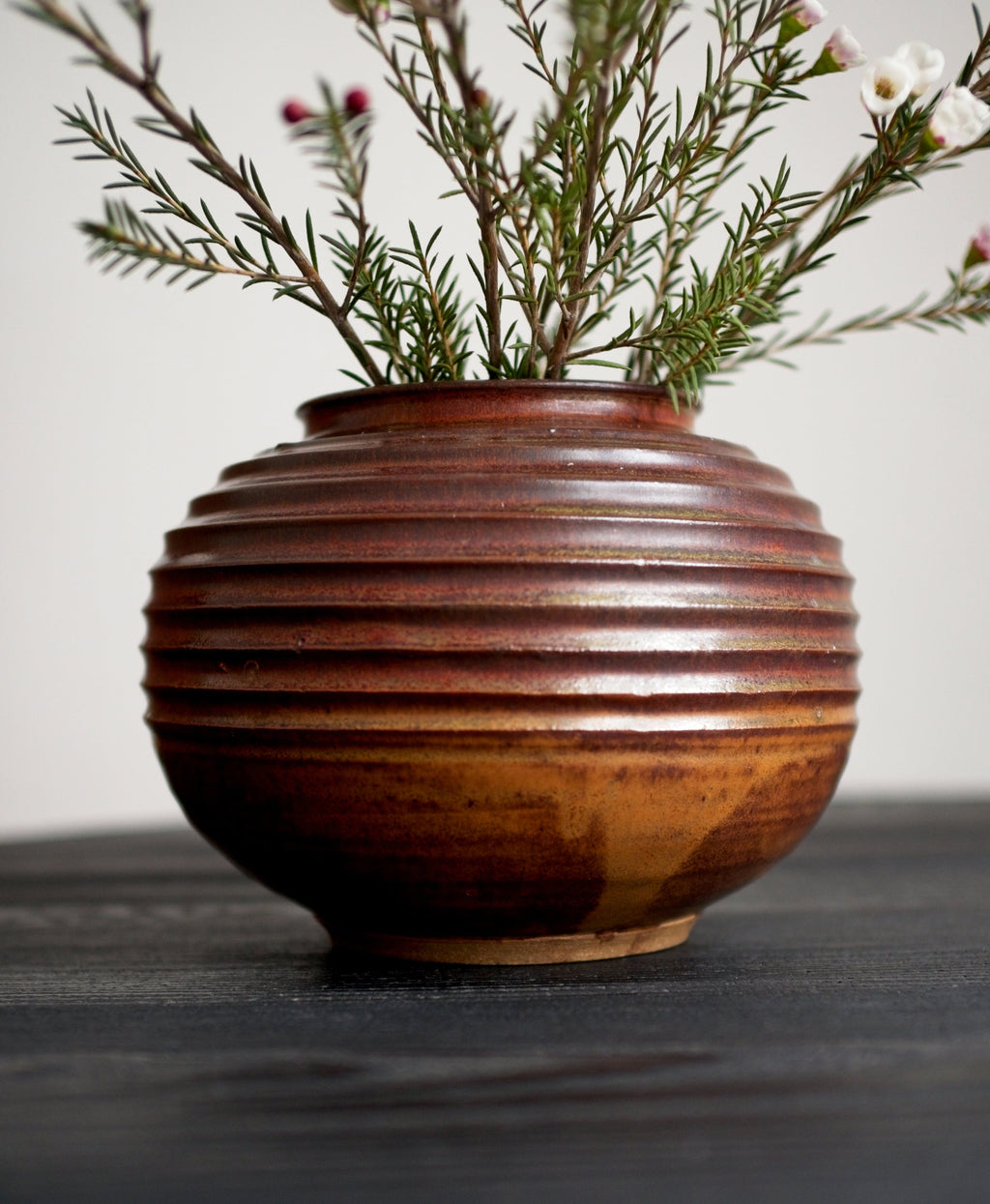 A round mid century modern brutalist style studio pottery ceramic vase with a reddish brown drip glaze and ribbed texture holding greenery on a dark surface.