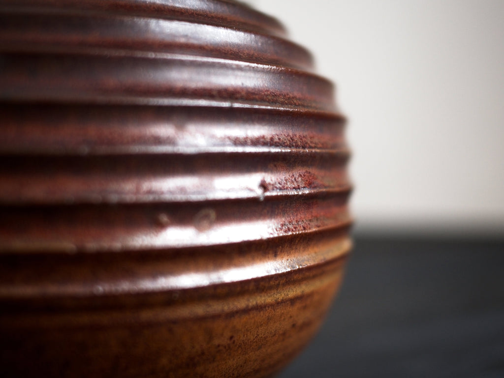A profile close-up of the carved ridges of reddish brown glazed vintage mid century modern spherically shaped studio pottery ceramic vase on a dark surface.