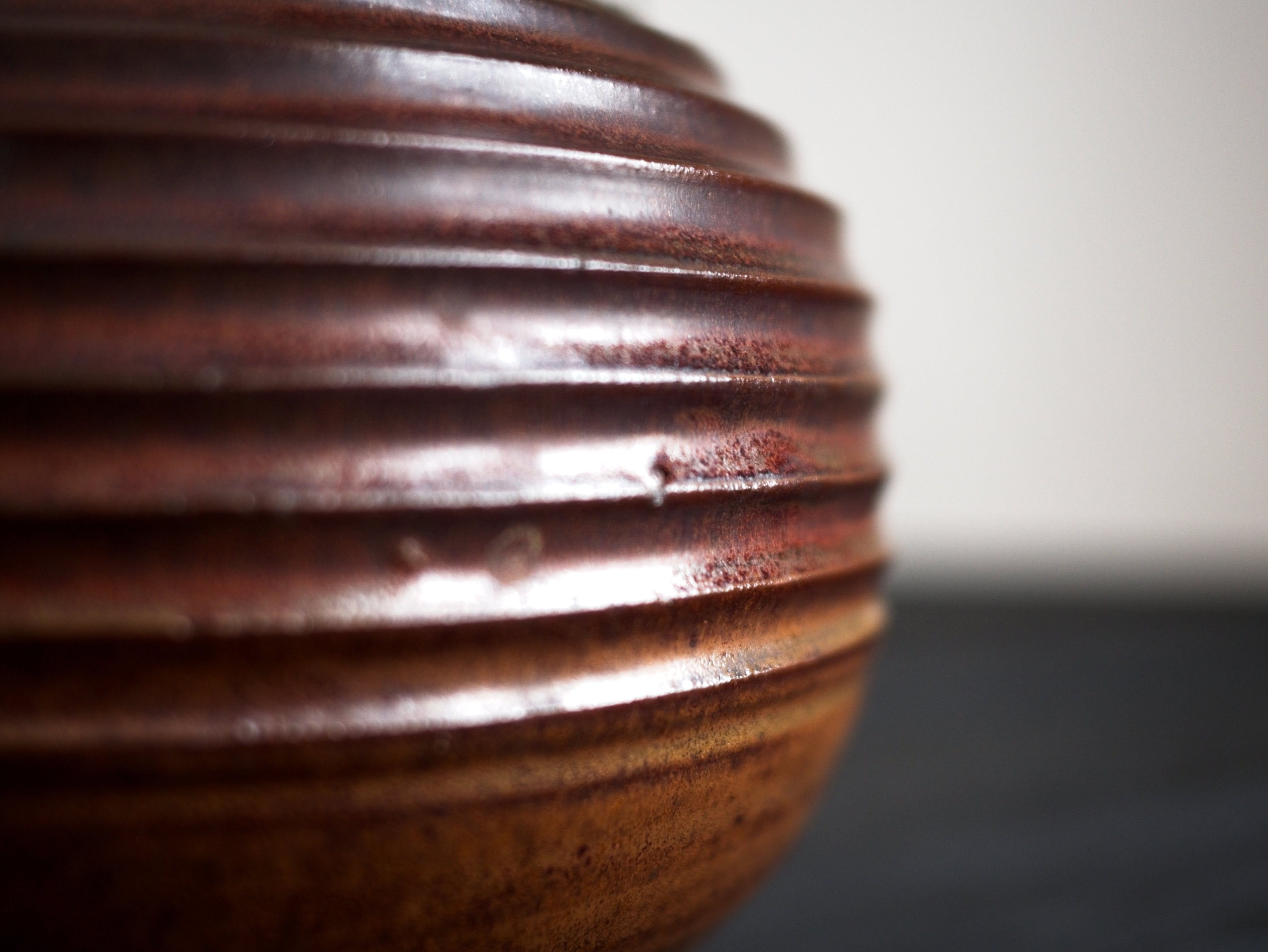A profile close-up of the carved ridges of reddish brown glazed vintage mid century modern spherically shaped studio pottery ceramic vase on a dark surface.