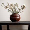 A vintage mid century modern art deco reddish brown ceramic studio pottery vase with an orange drip glaze containing flowers on a wooden table against a plain background.