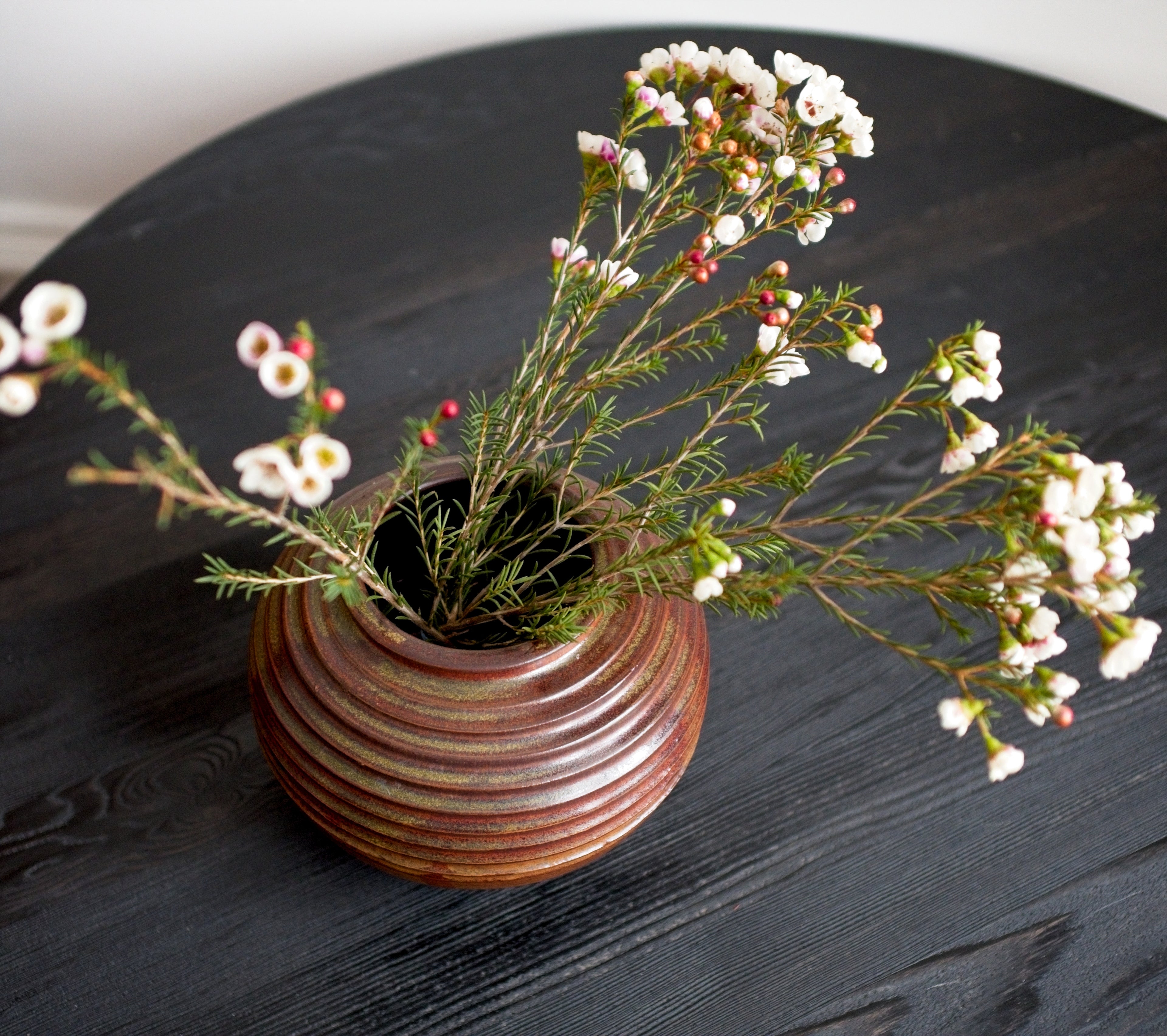 Ceramic brown vase with striped pattern containing small white flowers on a black wooden surface.