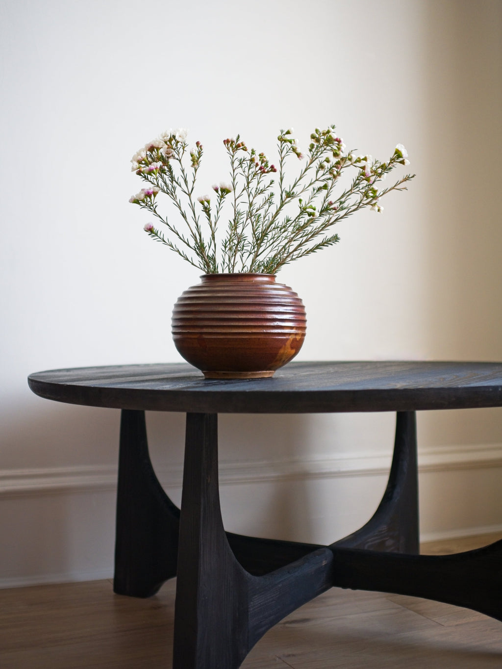 A vintage mid century modern spherical studio pottery style ceramic vase with a rustic brown glaze containing dried flowers on a dark wooden table against a plain wall.