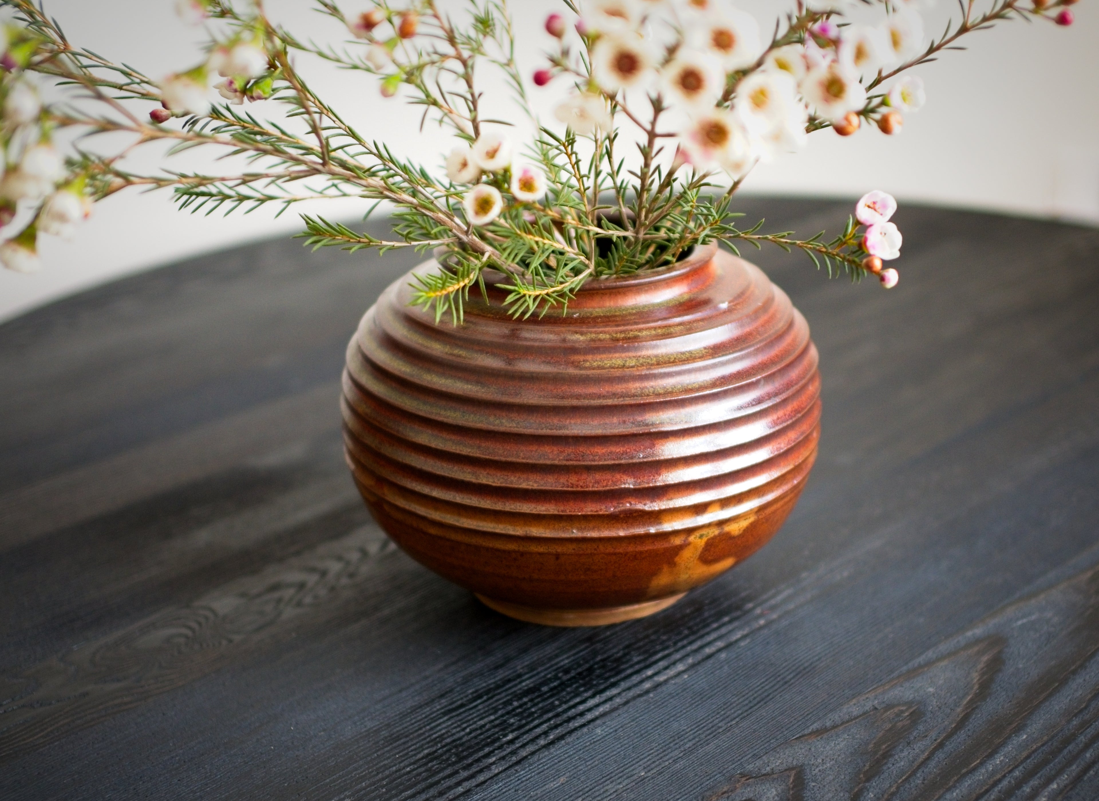Close up of a brown ribbed ceramic vase with small white flowers on a dark wooden surface.