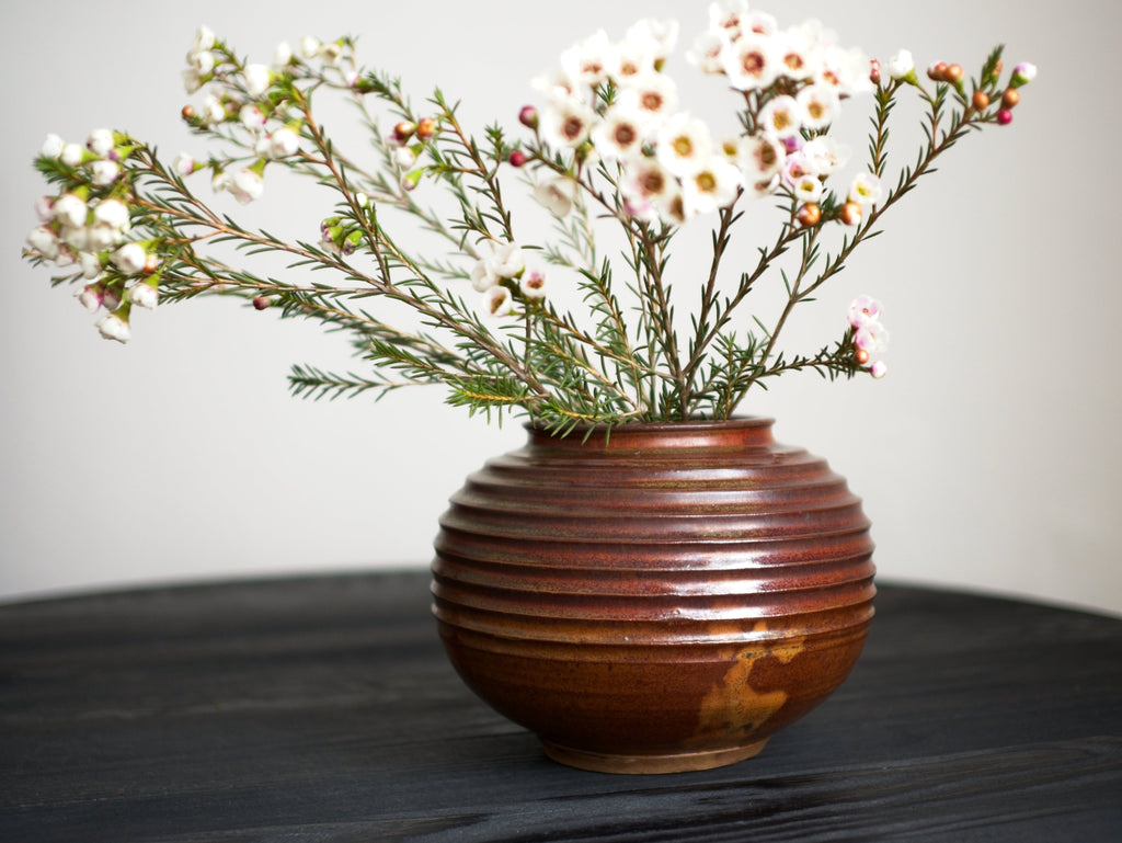 A vintage art deco spherical ribbed ceramic studio pottery vase with a reddish brown drip glaze containing small white flowers on a dark surface and light background.