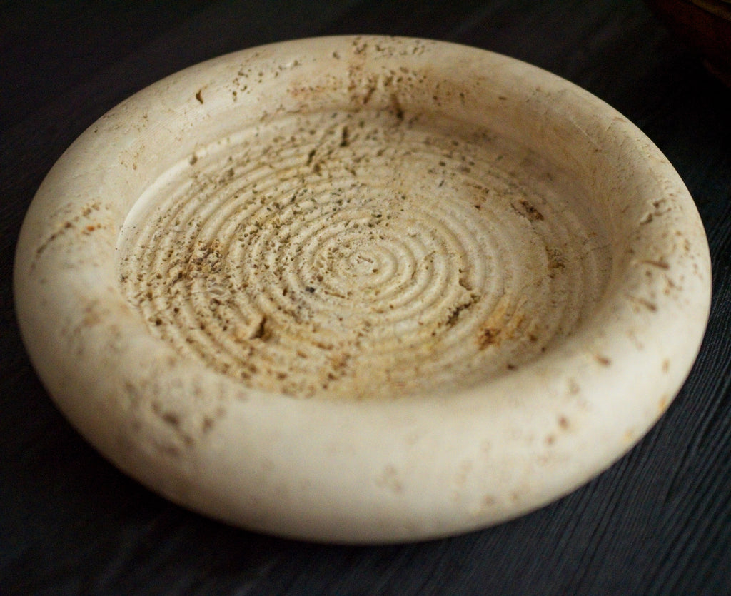 A close up of a 1970s vintage Fratelli Mannelli travertine vide poche on a black wood surface. The mid century modern travertine dish is hand carved with engraved circular ridge work inside, a wide sculptural rim and crackled texture throughout and is an beautiful example of classic retro Italian home decor that would suit modern vintage, retro, minimalist, Hollywood Regency, modernist or art deco inspired home interior schemes.