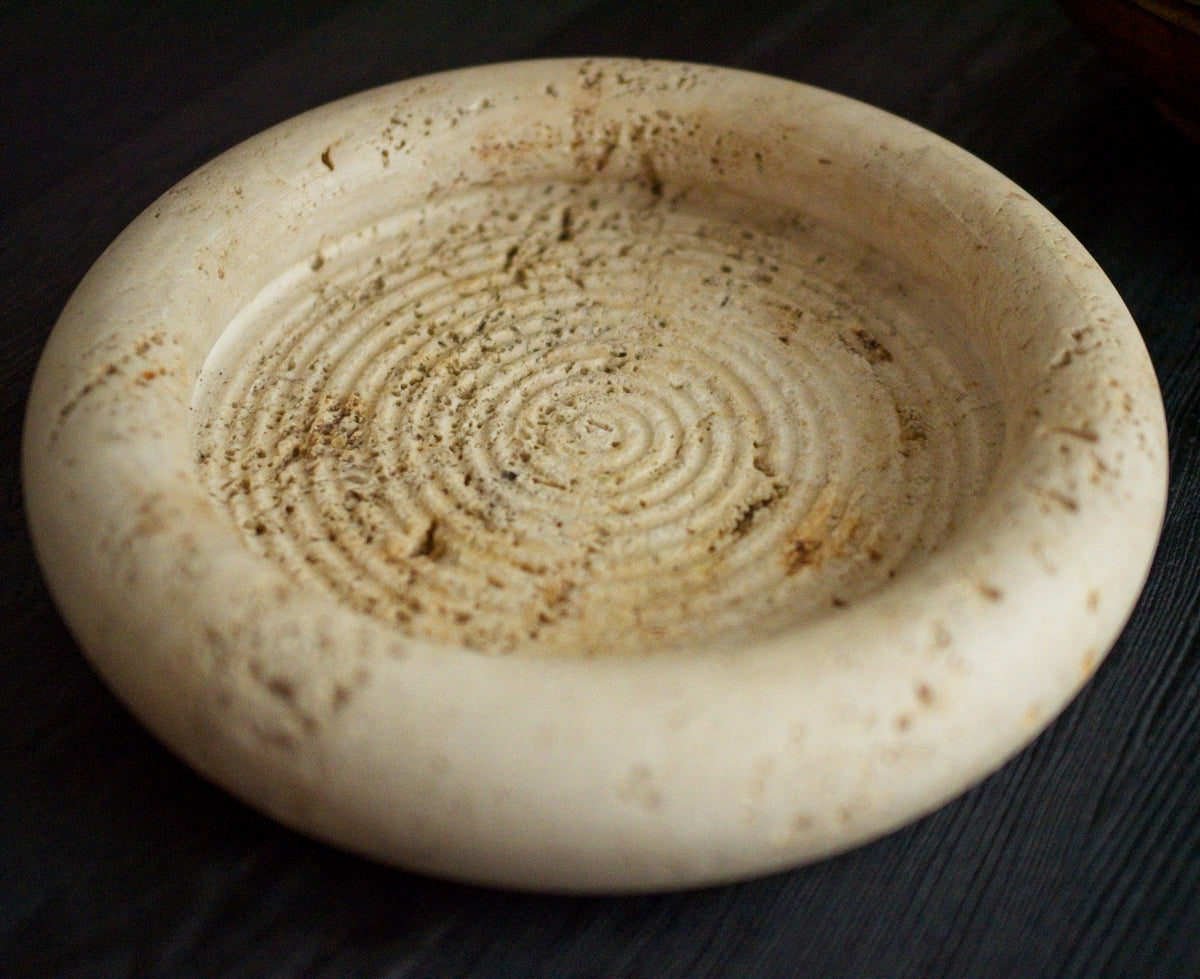 A close up of a 1970s vintage Fratelli Mannelli travertine vide poche on a black wood surface. The mid century modern travertine dish is hand carved with engraved circular ridge work inside, a wide sculptural rim and crackled texture throughout and is an beautiful example of classic retro Italian home decor that would suit modern vintage, retro, minimalist, Hollywood Regency, modernist or art deco inspired home interior schemes.