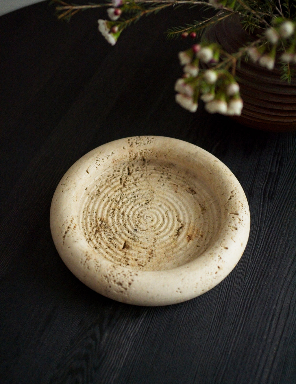 A 1970s vintage Fratelli Mannelli travertine vide poche next to a brutalist globe vase containing small white rustic flowers on a black wood surface. This round mid century modern travertine dish is hand carved with engraved circular ridge work inside, a wide sculptural rim and crackled texture throughout and is an beautiful example of classic retro Italian home decor that would suit modern vintage, retro, minimalist, Hollywood Regency, modernist or art deco inspired home interior schemes.