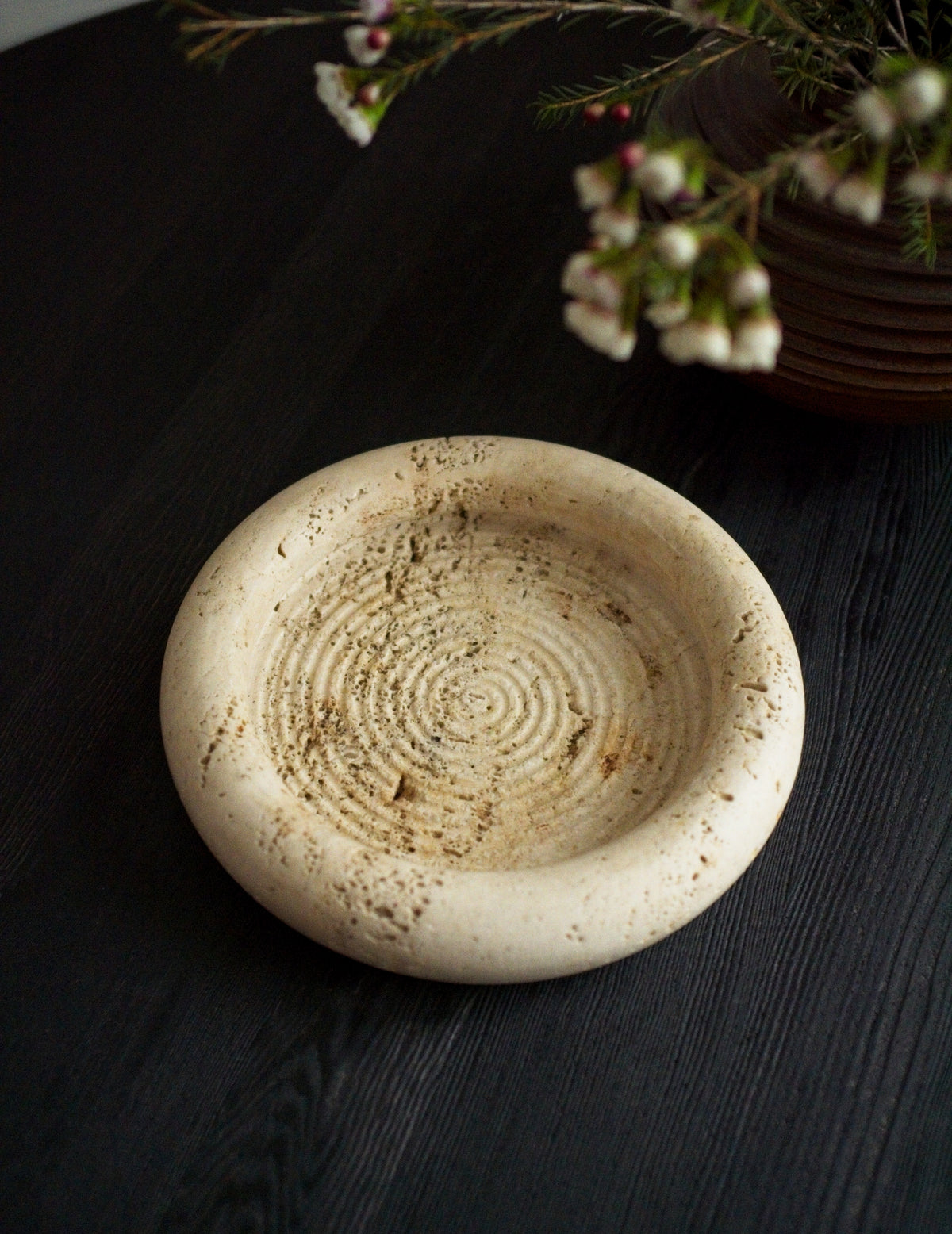 A 1970s vintage Fratelli Mannelli travertine vide poche next to a brutalist globe vase containing small white rustic flowers on a black wood surface. This round mid century modern travertine dish is hand carved with engraved circular ridge work inside, a wide sculptural rim and crackled texture throughout and is an beautiful example of classic retro Italian home decor that would suit modern vintage, retro, minimalist, Hollywood Regency, modernist or art deco inspired home interior schemes.