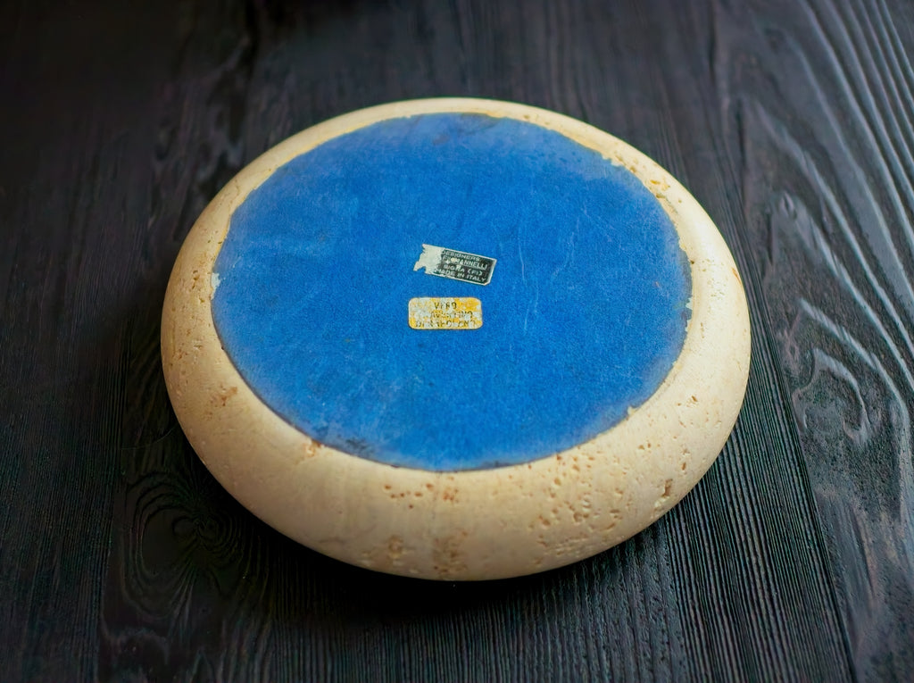 Underneath of a round ceramic item with a blue backing featuring black and gold brand labels in the centre on a black wooden surface.