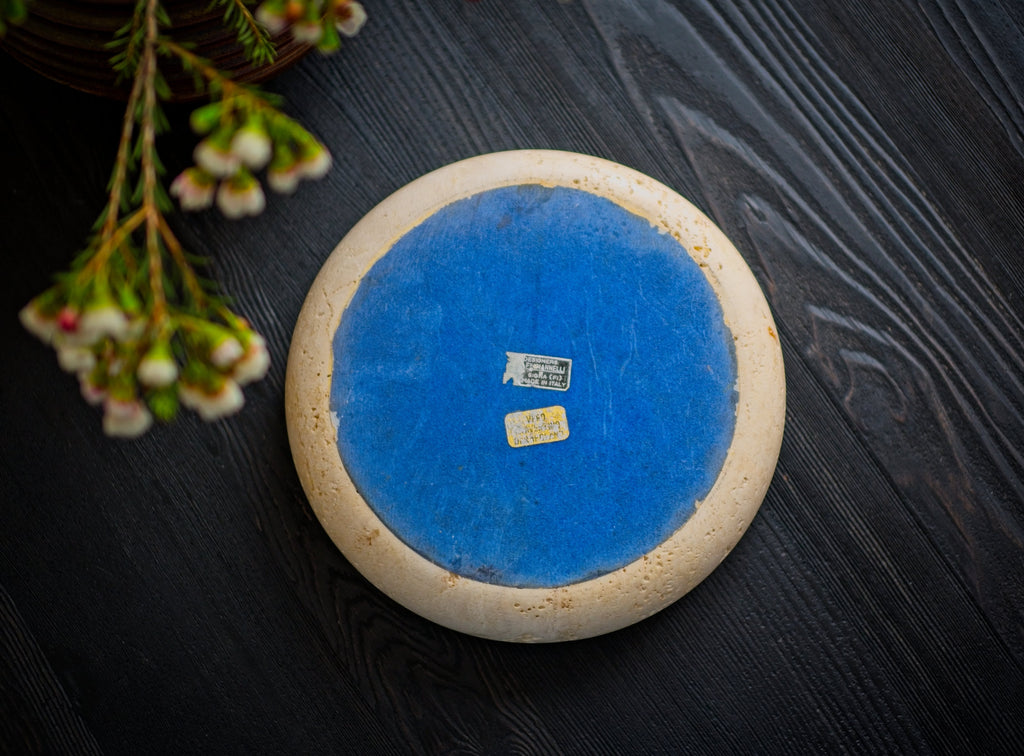 The bottom of a 1970s mid century modern vintage Fratelli Mannelli travertine round stone dish with blue fabric backing featuring two authentic designer labels on a dark wooden surface.