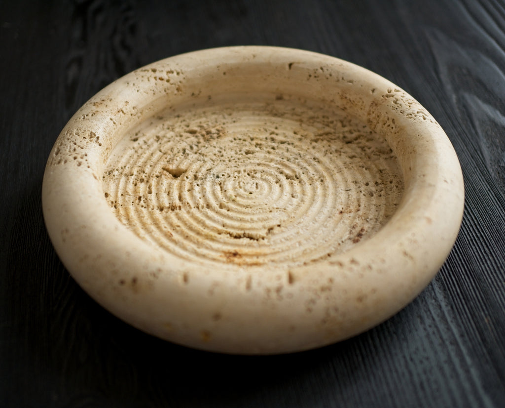 Close up of a round beige stone dish with concentric circular ridged pattern on a black wooden surface.
