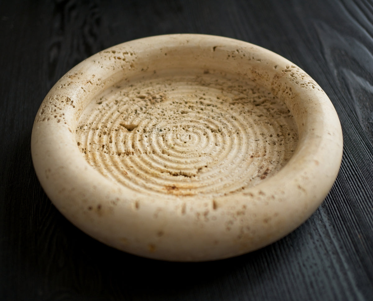 A 1970s vintage Fratelli Mannelli travertine vide poche on a black wood surface. The mid century modern travertine dish is hand carved with engraved circular ridge work inside, a wide sculptural rim and crackled texture throughout and is an beautiful example of classic retro Italian home decor that would suit modern vintage, retro, minimalist, Hollywood Regency, modernist or art deco inspired home interior schemes.