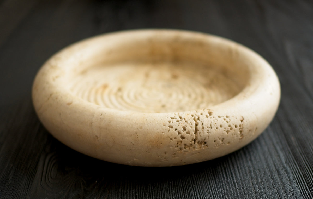 A close up of the rim of a 1970s vintage Fratelli Mannelli travertine vide poche on a black wood surface. The mid century modern travertine dish is hand carved with engraved circular ridge work inside, a wide sculptural rim and crackled texture throughout and is an beautiful example of classic retro Italian home decor that would suit modern vintage, retro, minimalist, Hollywood Regency, modernist or art deco inspired home interior schemes.