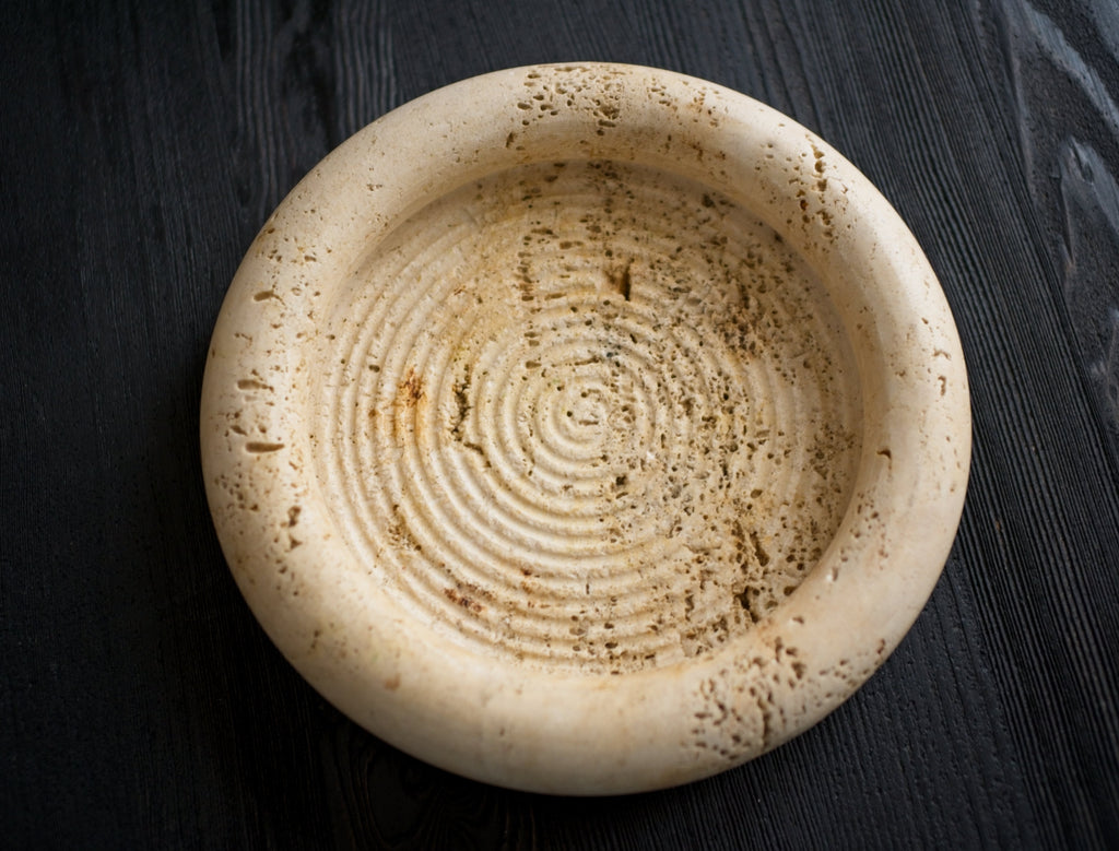A chic 1970s mid century modern vintage Fratelli Mannelli solid travertine circular beige stone dish with textured surface on a dark wooden background from above.