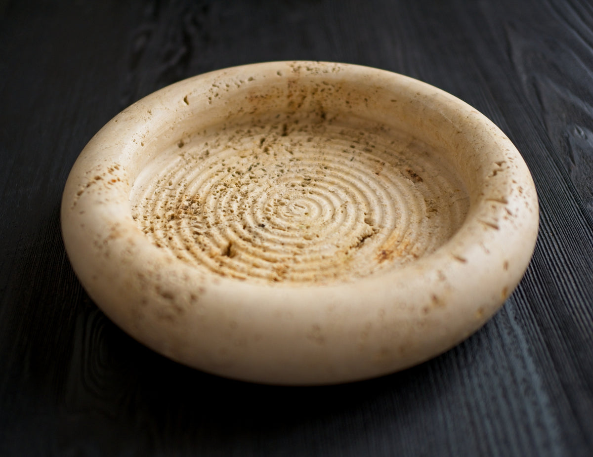 A 1970s vintage Fratelli Mannelli travertine vide poche on a black wood surface. The mid century modern travertine dish is hand carved with engraved circular ridge work inside, a wide sculptural rim and crackled texture throughout and is an beautiful example of classic retro Italian home decor that would suit modern vintage, retro, minimalist, Hollywood Regency, modernist or art deco inspired home interior schemes.