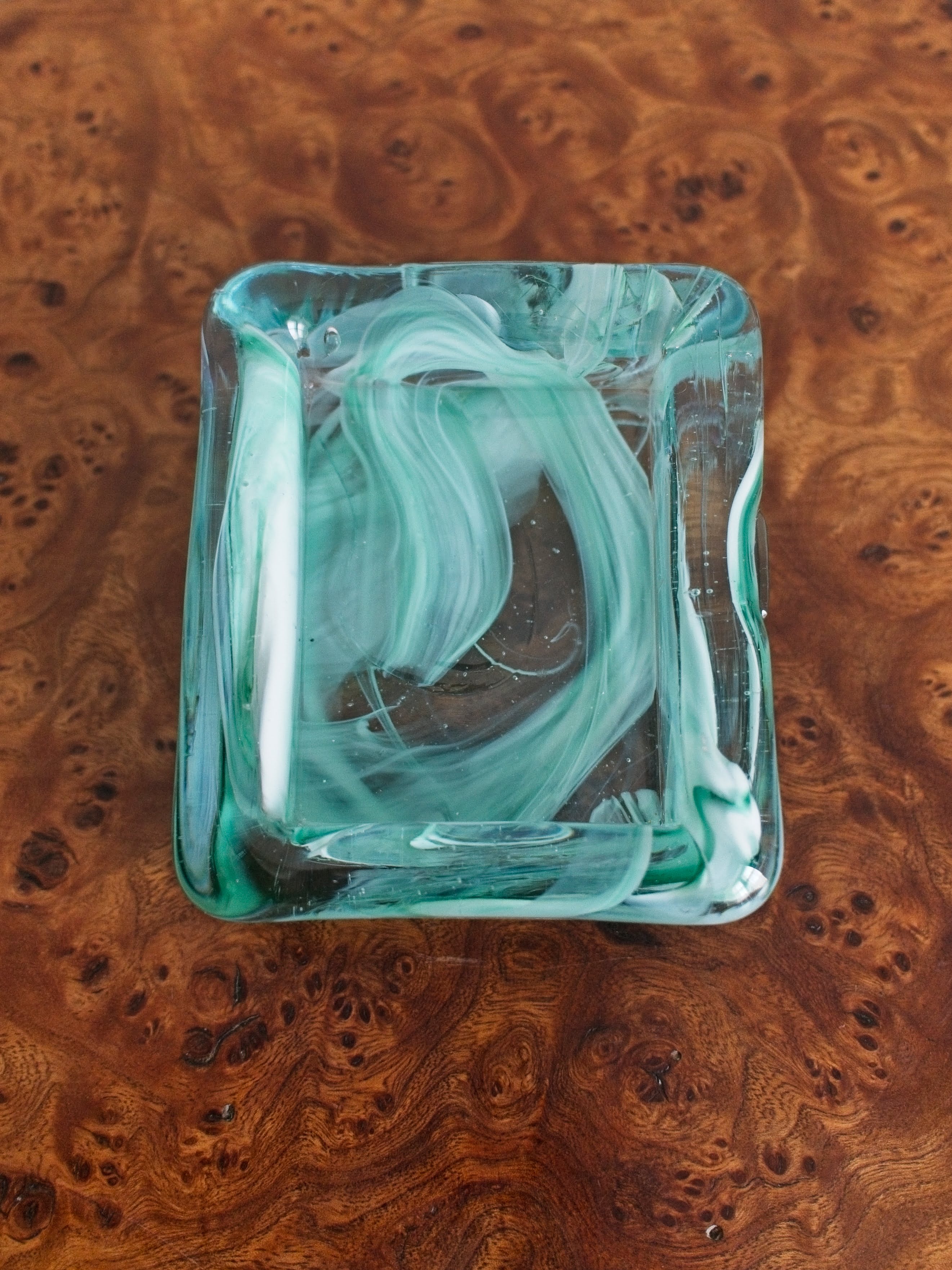 A small rectangular 1970s mid century modern glass trinket dish with green and white marble swirls similar to Murano glass containing a variety of silver jewellery set on a vintage burr wood table.