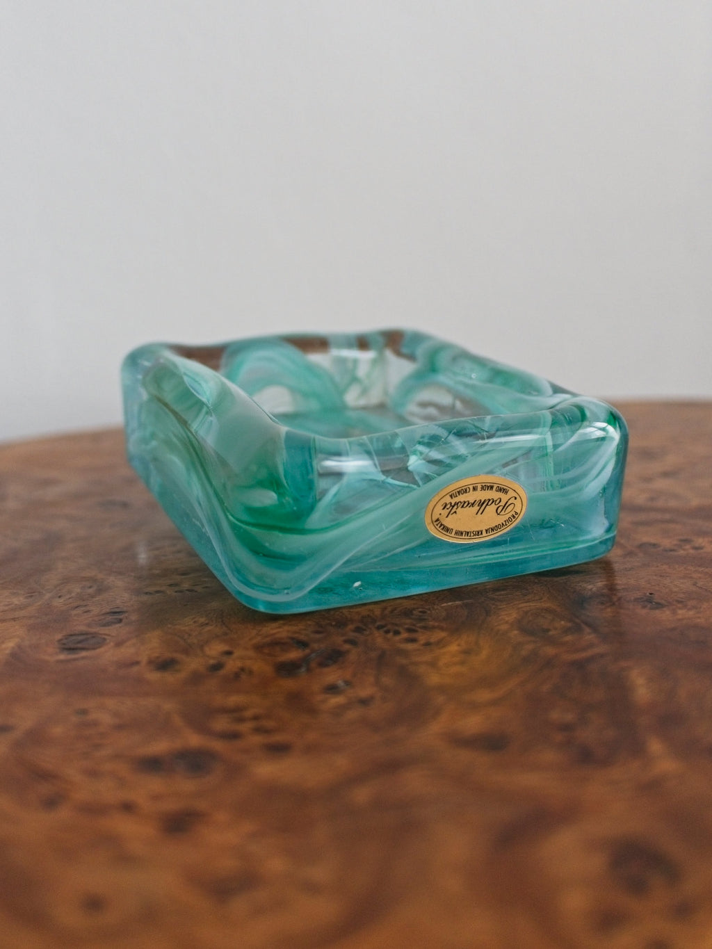 A small 1970s mid century modern glass trinket dish with green and white marble swirls similar to Murano glass with a gold sticker saying 'Podhraski' on a vintage burr wood table.