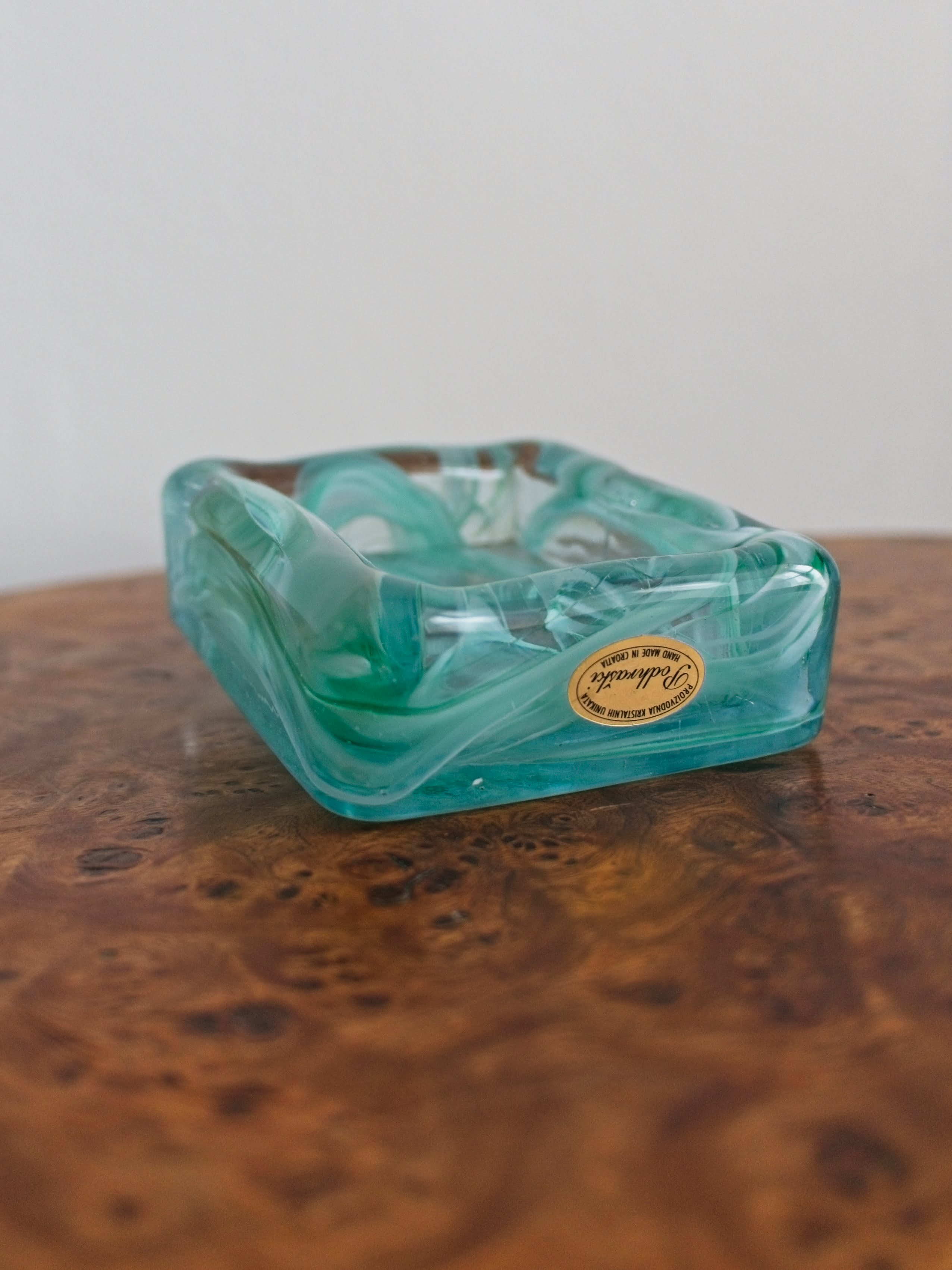 A small 1970s mid century modern glass trinket dish with green and white marble swirls similar to Murano glass with a gold sticker saying 'Podhraski' on a vintage burr wood table.