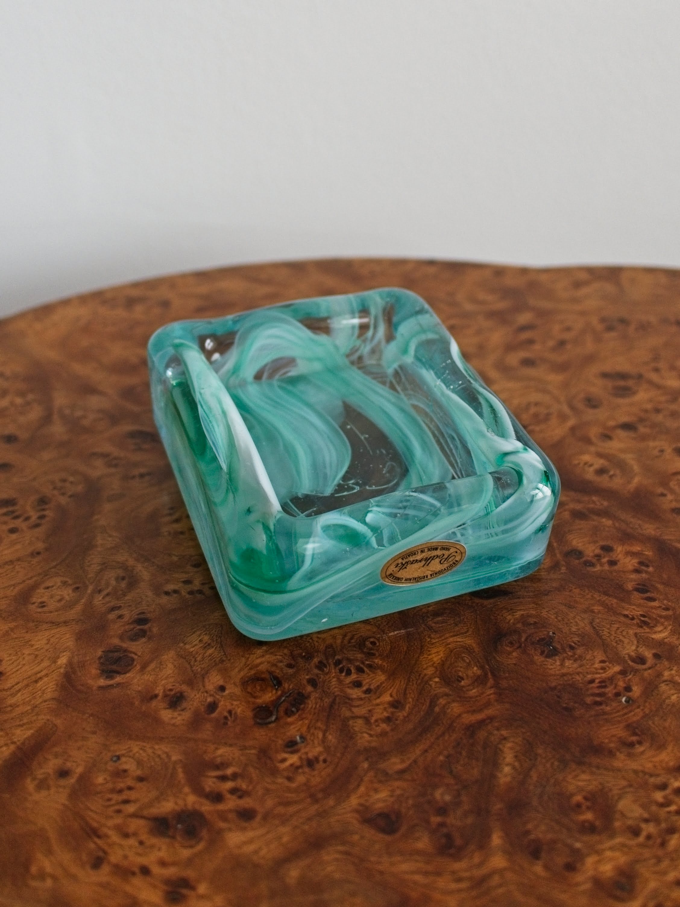 A small 1970s retro mid century modern glass trinket dish with green and white marble swirls similar to Murano glass with a gold sticker saying 'Podhraski' on a vintage burr wood table.