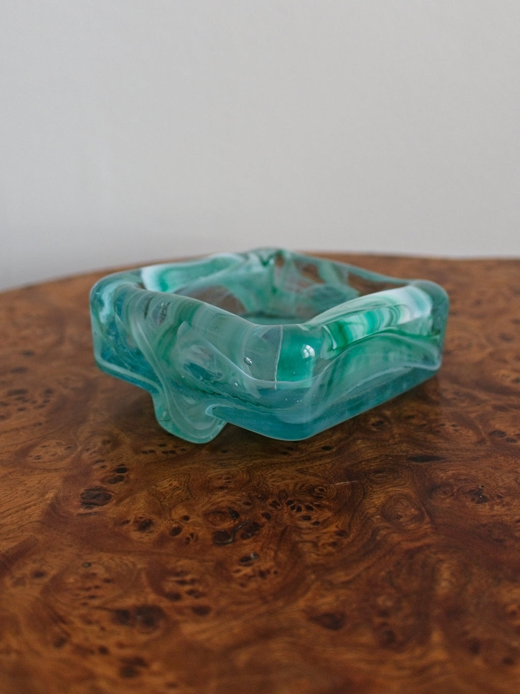 The back of a small 1970s mid century modern glass trinket dish with green and white marble swirls similar to Murano glass on a vintage burr wood table.