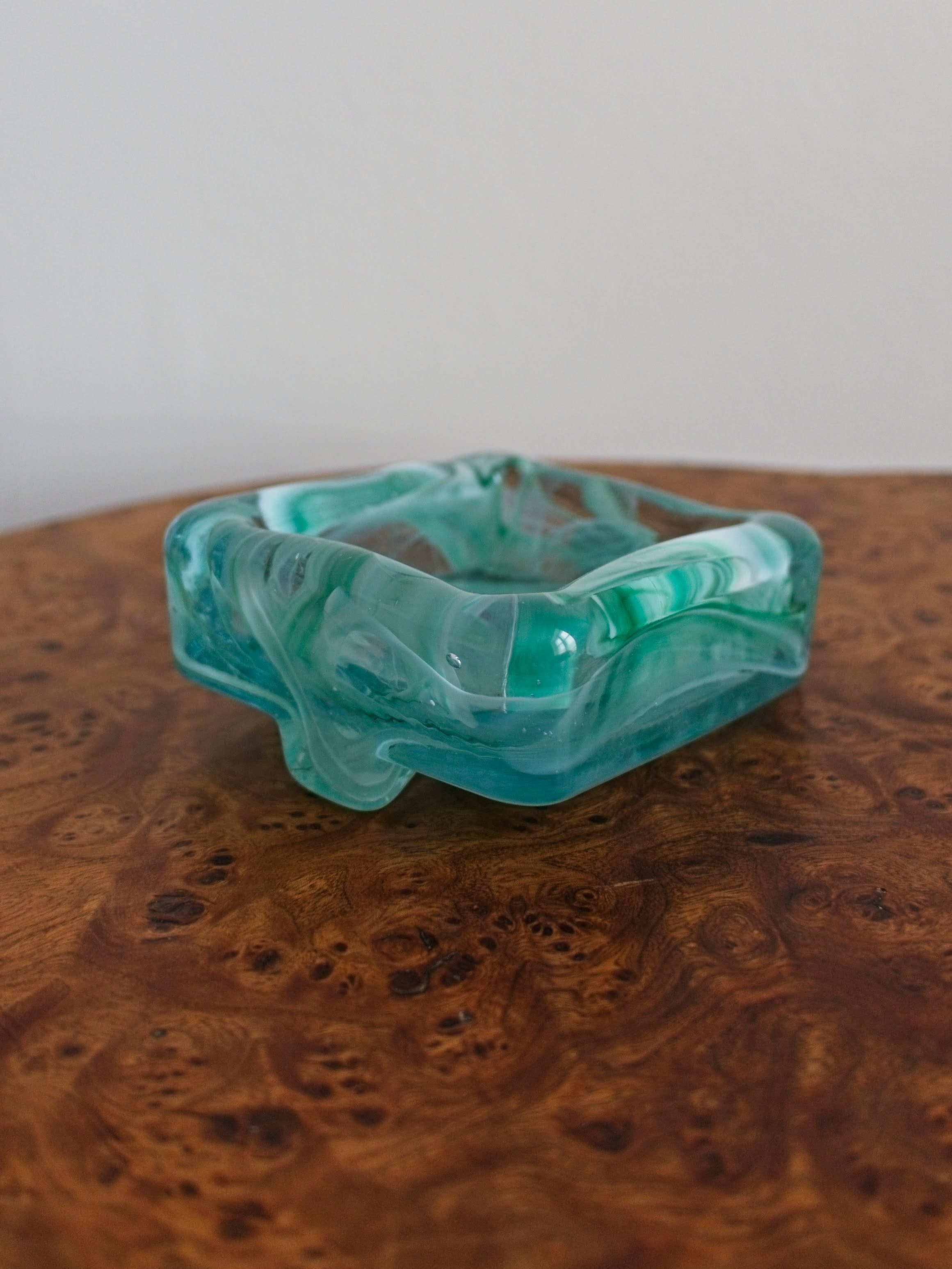 The back of a small 1970s mid century modern glass trinket dish with green and white marble swirls similar to Murano glass on a vintage burr wood table.