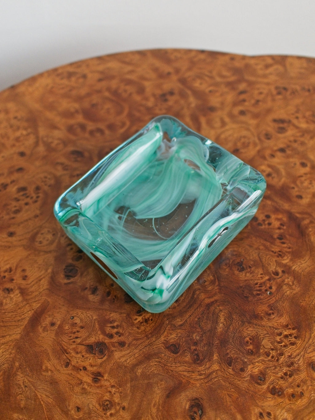 A small 1970s mid century modern glass trinket dish with green and white marble swirls similar to Murano glass on a vintage burr wood table.
