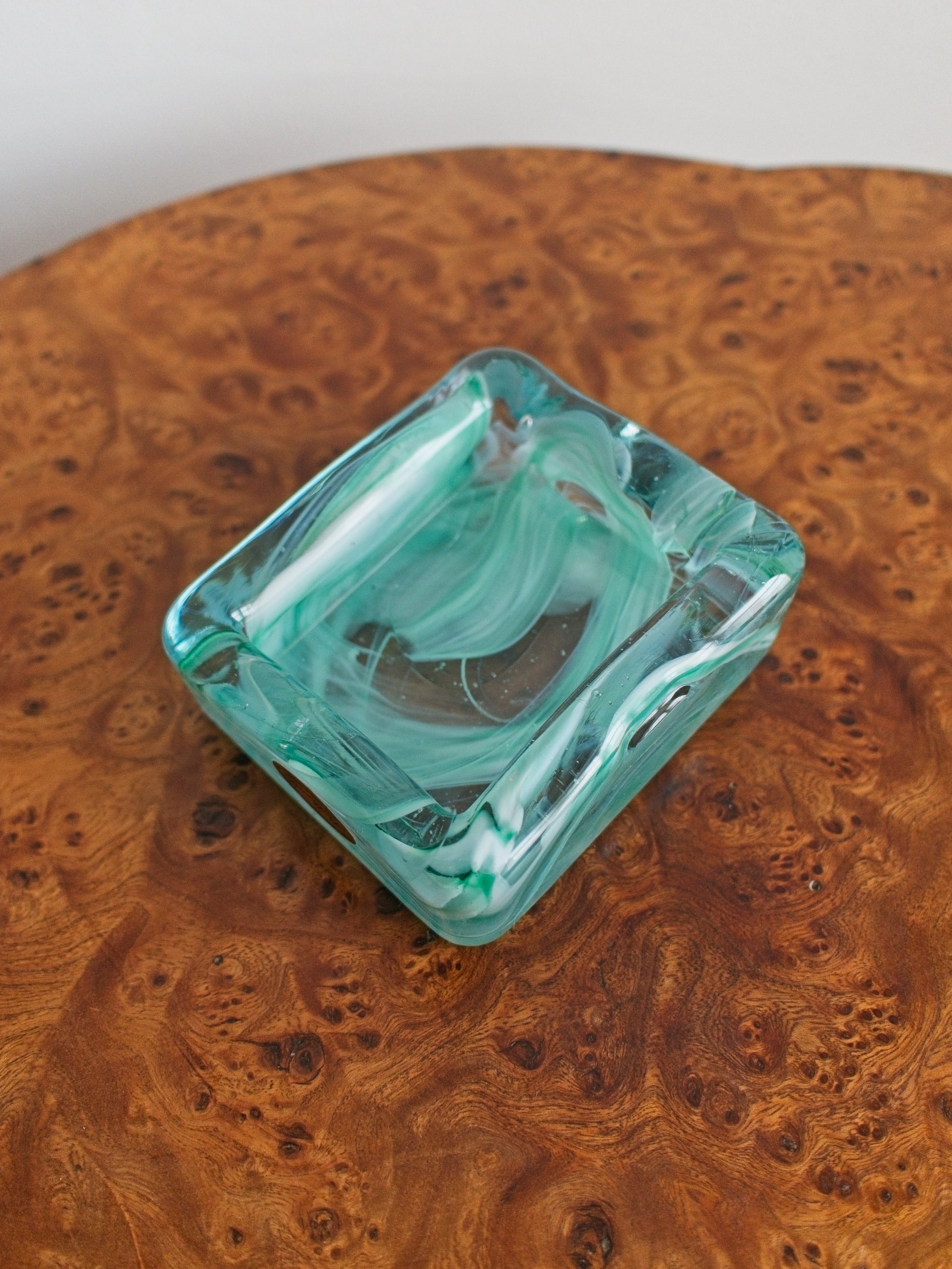 A small 1970s mid century modern glass trinket dish with green and white marble swirls similar to Murano glass on a vintage burr wood table.