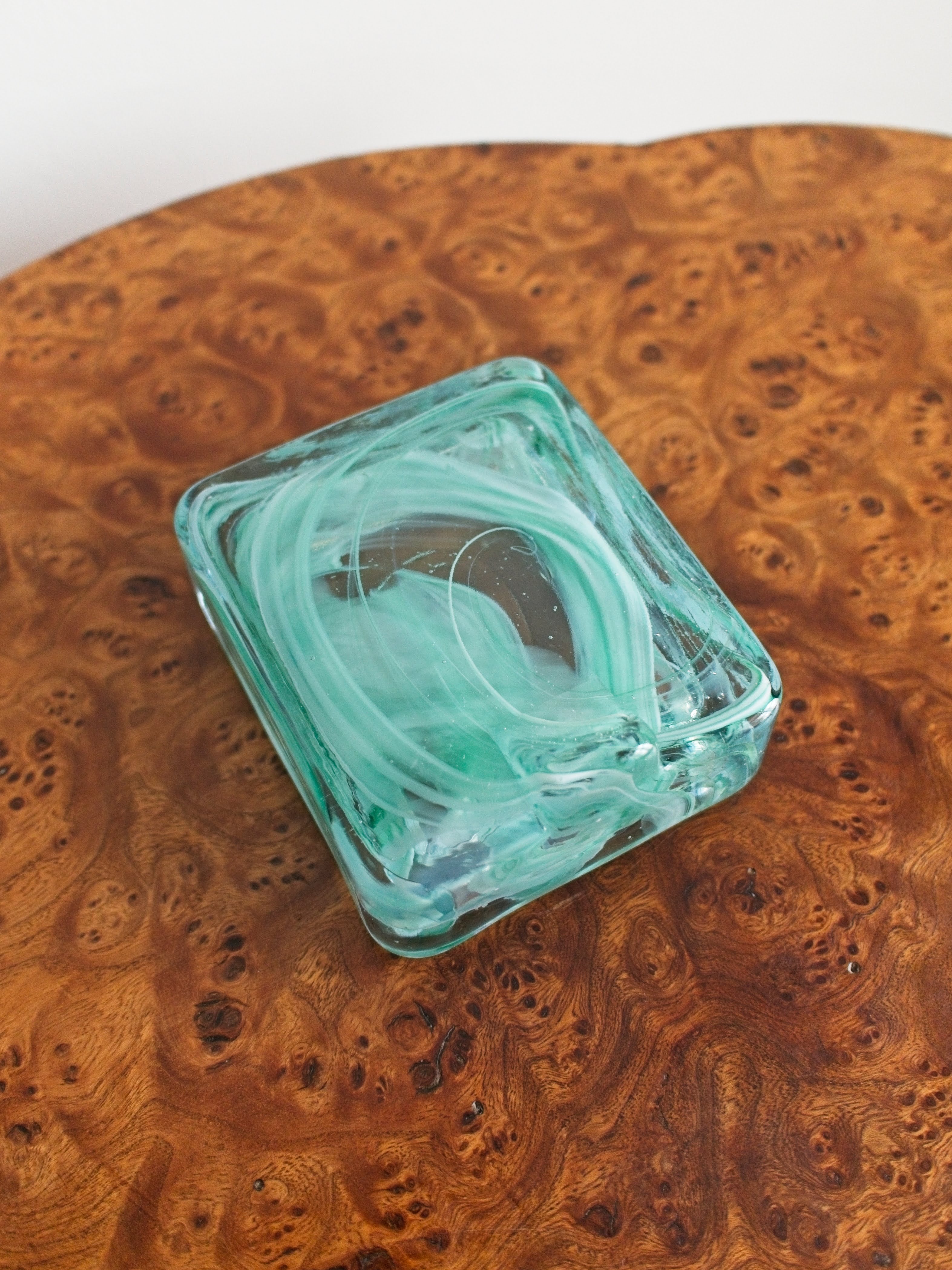 The bottom of a small 1970s mid century modern glass trinket dish with green and white marble swirls similar to Murano glass  on a vintage burr wood table.