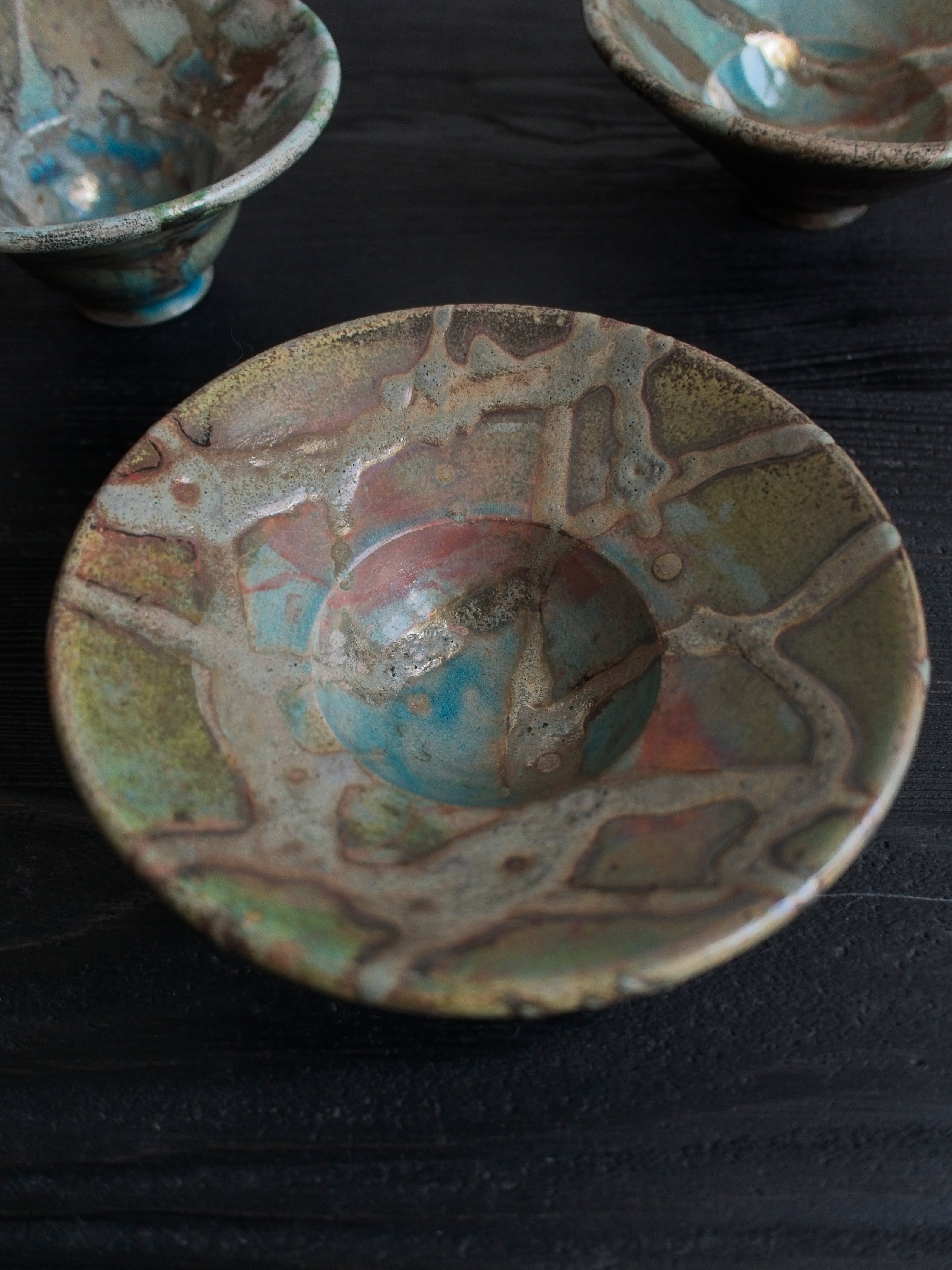 A close up of a green, blue and orange vintage Japanese handmade studio pottery décor bowl with abstract oxidised iridescent glaze patterns on a black wooden table with two similar bowls in the background.