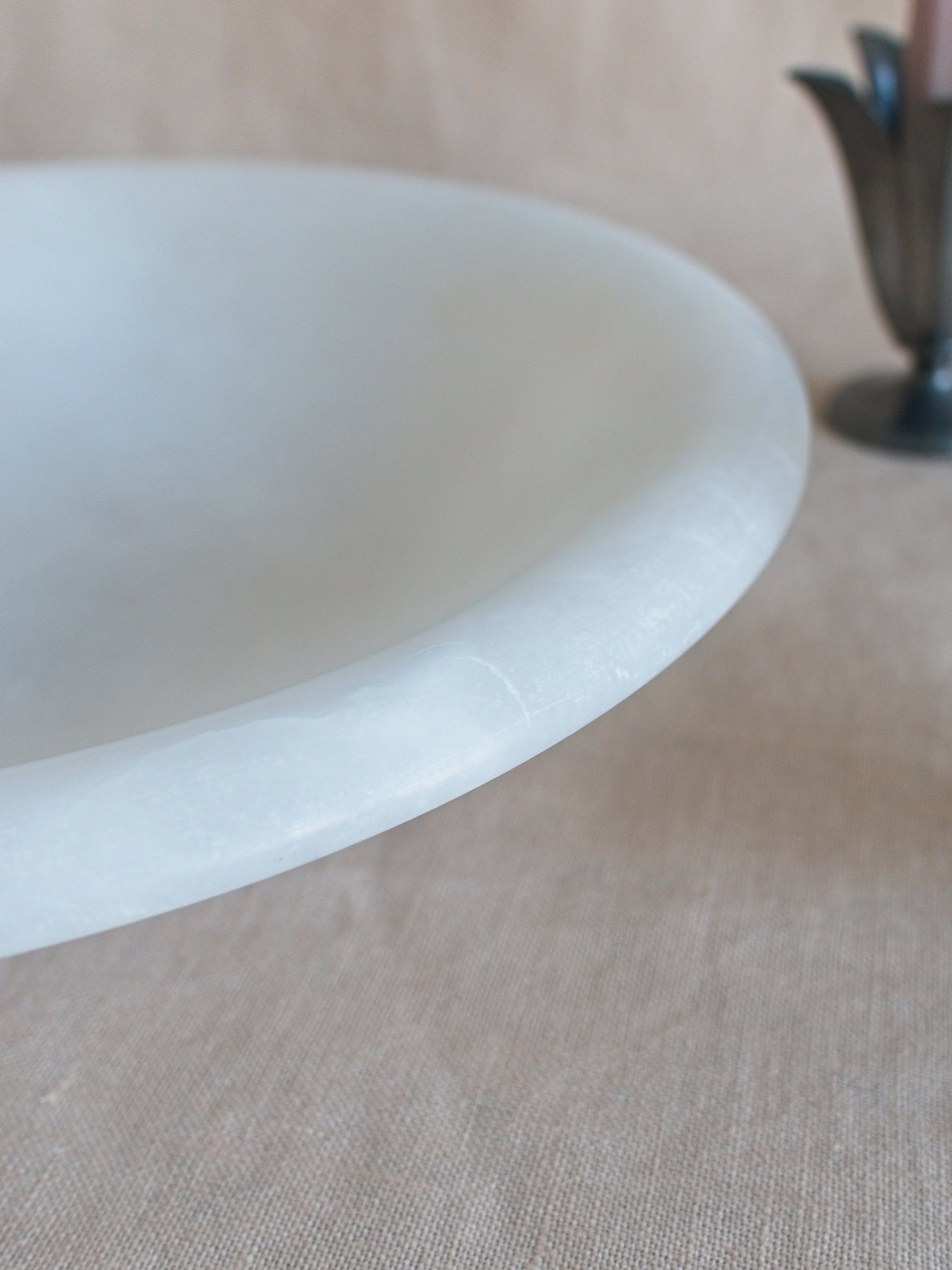 A close up showing the curved rim of a vintage art deco style white alabaster decorative bowl by Volterra, Italy, with a small white mark in front of a pewter tulip shaped 1930s candle holder by Just Andersen