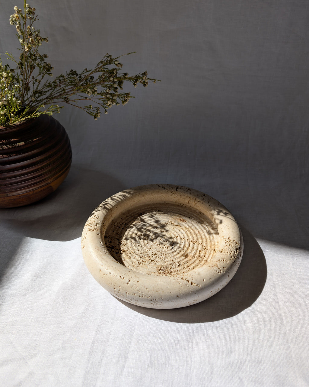 A vintage 1970s mid century modern Fratelli Mannelli travertine vide poche display dish on a white linen surface next to an art deco style brown ceramic globe vase with dried rustic flowers.
