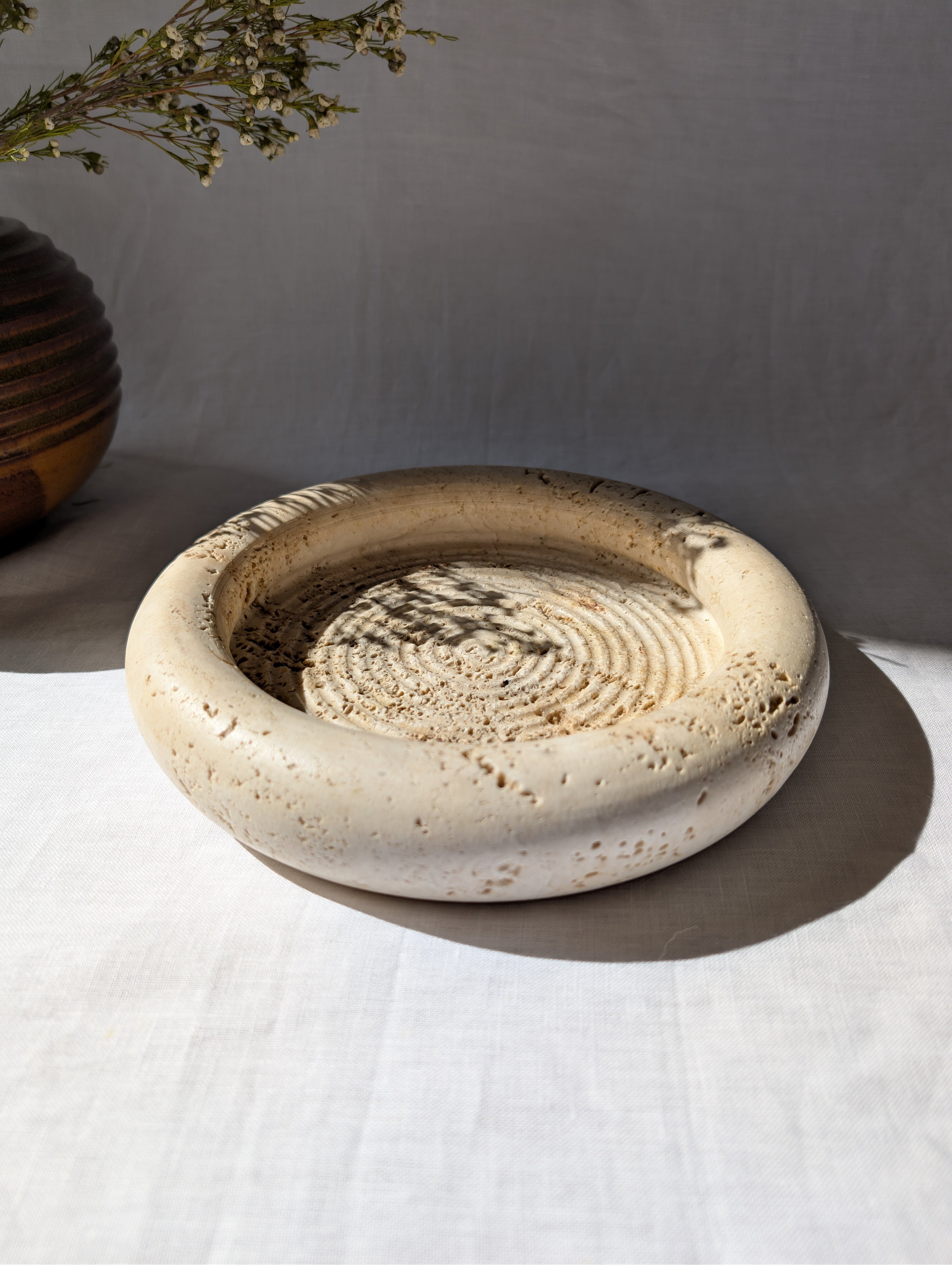 A close up of a vintage 1970s mid century modern Fratelli Mannelli travertine vide poche display dish on a white linen surface next to an art deco style brown ceramic globe vase with dried rustic flowers.