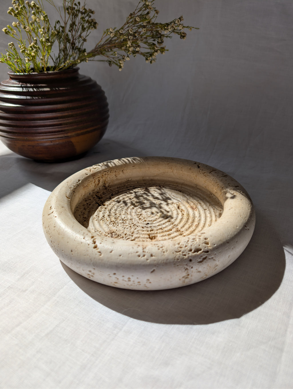 A vintage 1970s mid century modern Fratelli Mannelli travertine vide poche display dish on a white linen surface next to an art deco style brown ceramic globe vase with dried rustic flowers.