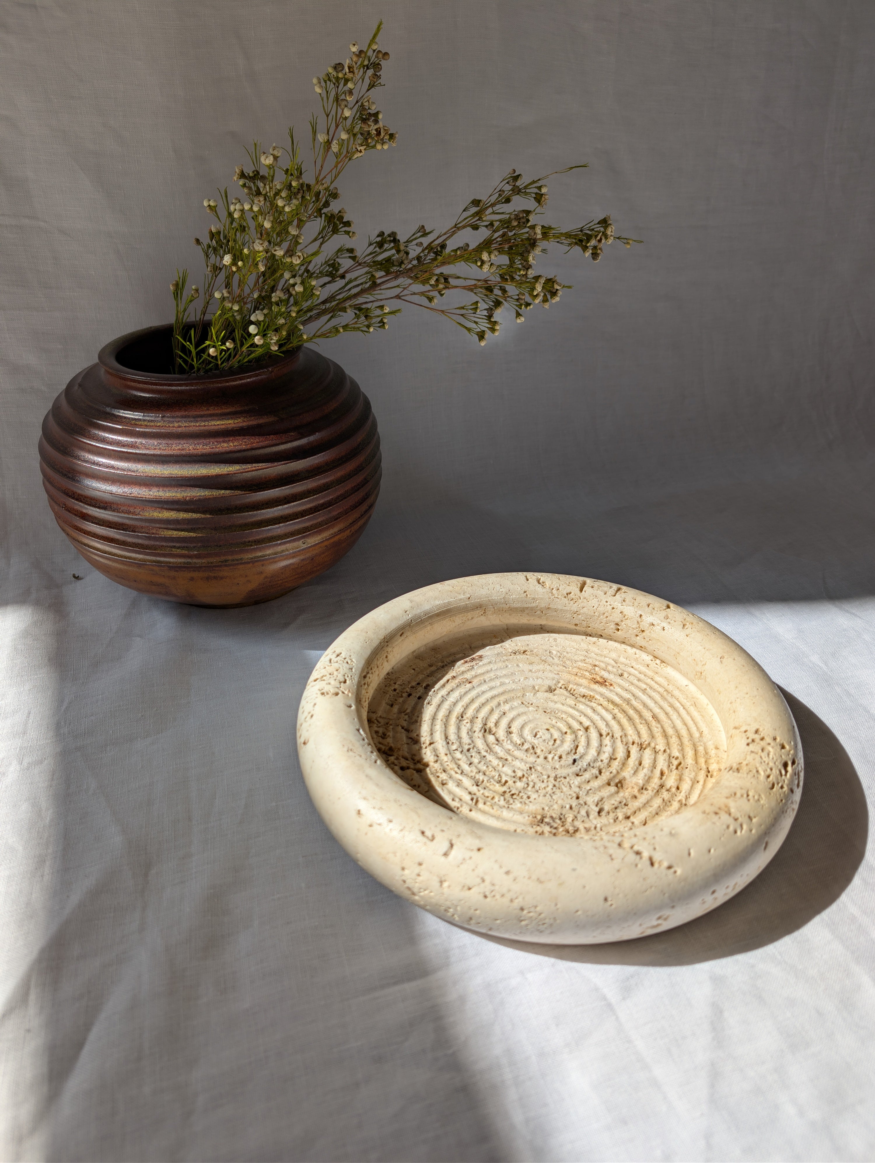 A vintage 1970s mid century modern Fratelli Mannelli travertine vide poche display dish on a white linen surface next to an art deco style brown ceramic globe vase with dried rustic flowers.