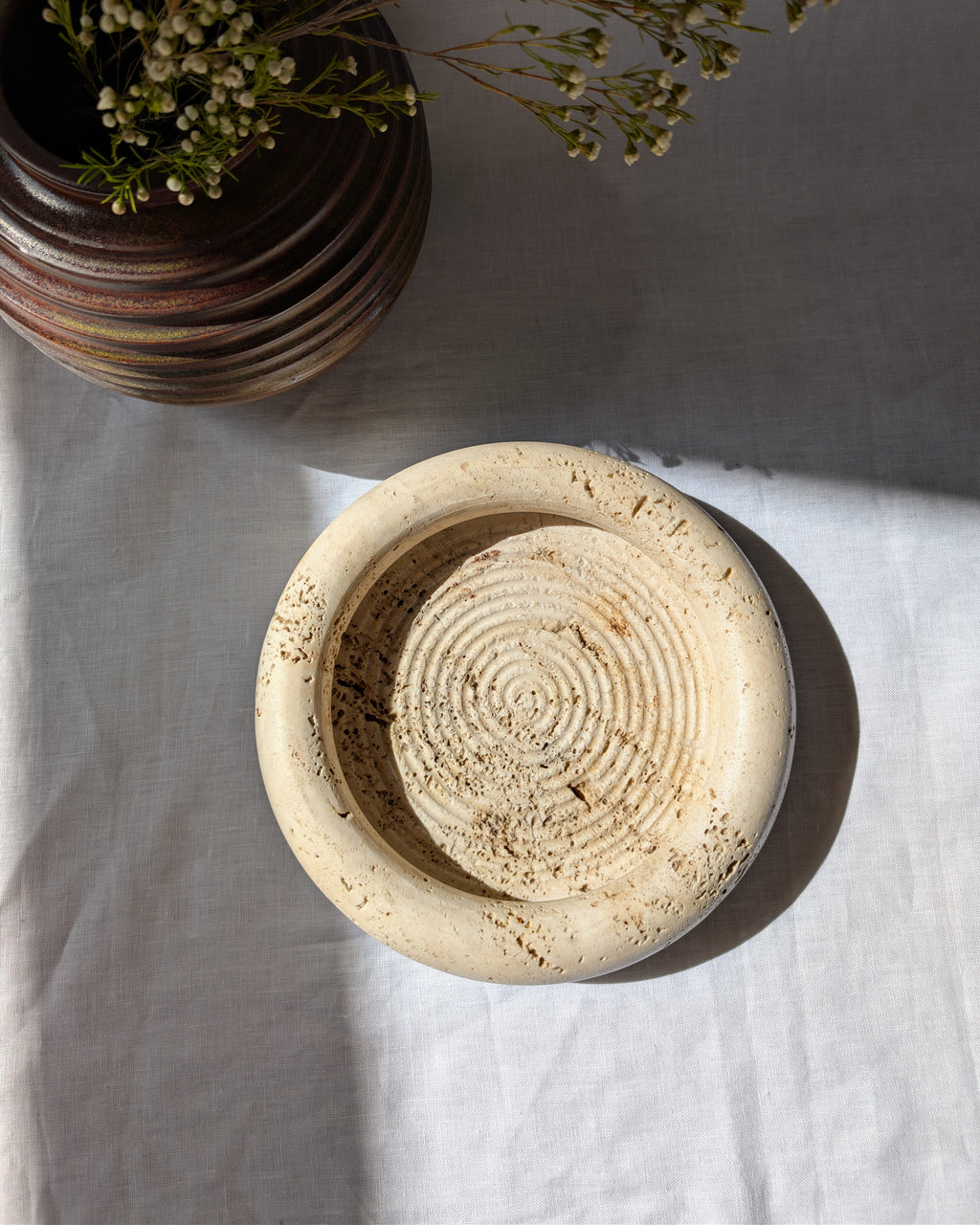 A vintage 1970s mid century modern Fratelli Mannelli travertine vide poche display dish on a white linen surface next to an art deco style brown ceramic globe vase with dried rustic flowers.