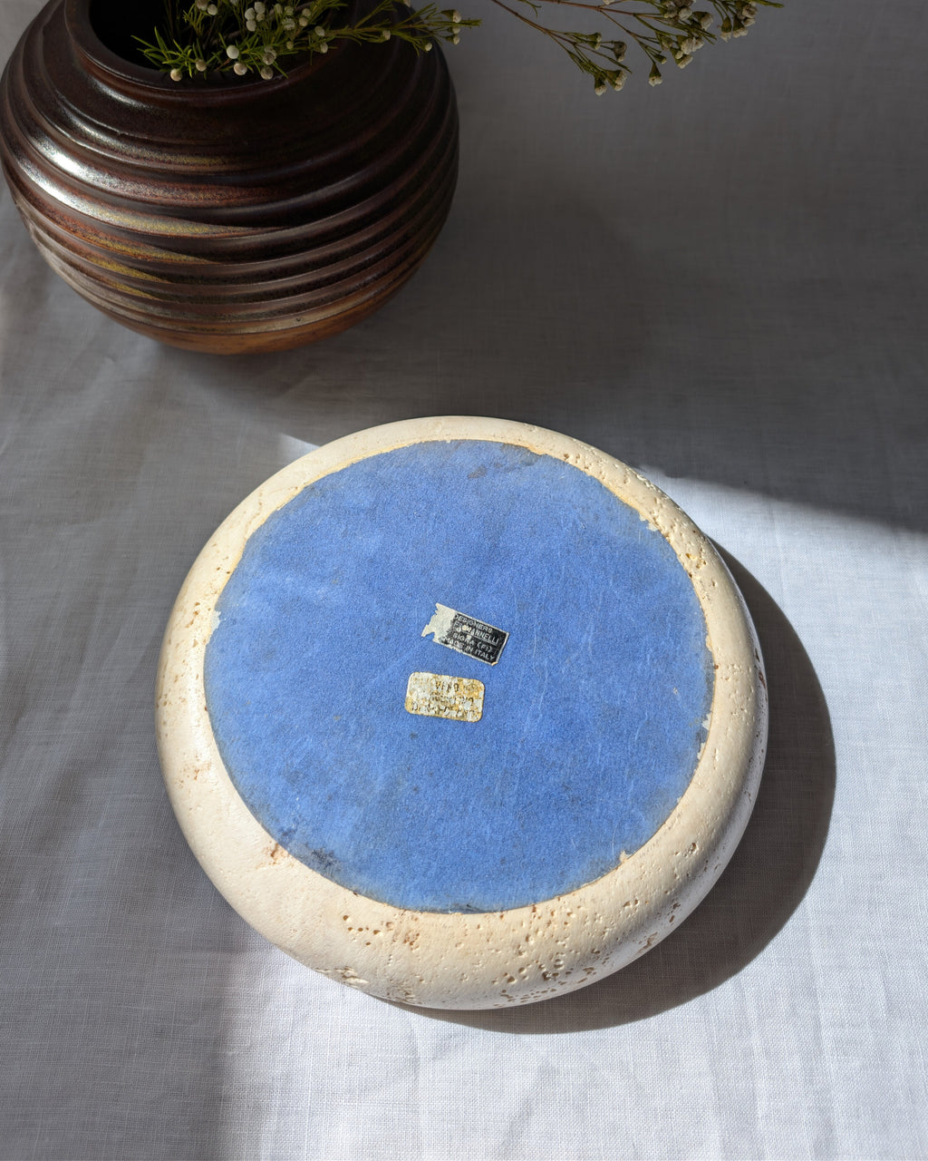 The base of a 1970s mid century modern vintage Fratelli Mannelli travertine circular stone display dish with blue fabric backing featuring two black and gold authenticity labels on a white linen surface next to a brutalist art deco style brown ceramic globe vase with small rustic flowers.