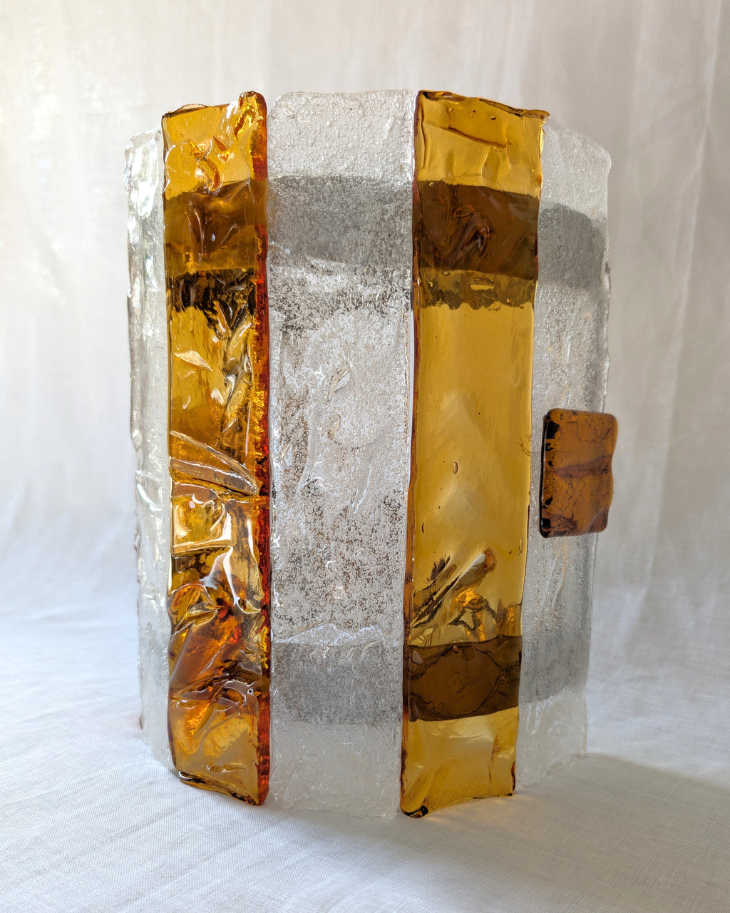 A close up of a vintage mid century modern 1970s Carlo Nason for Mazzega rectangular wall sconce with textured amber and clear striped Murano glass panels on white linen surface.